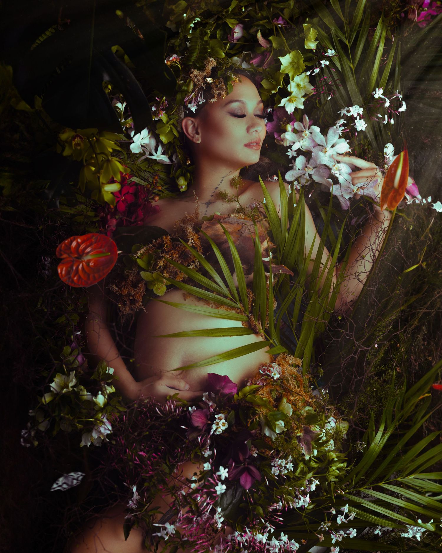 A figure lies among tropical foliage and flowers in a dreamy nature-inspired portrait with dramatic lighting and colors.