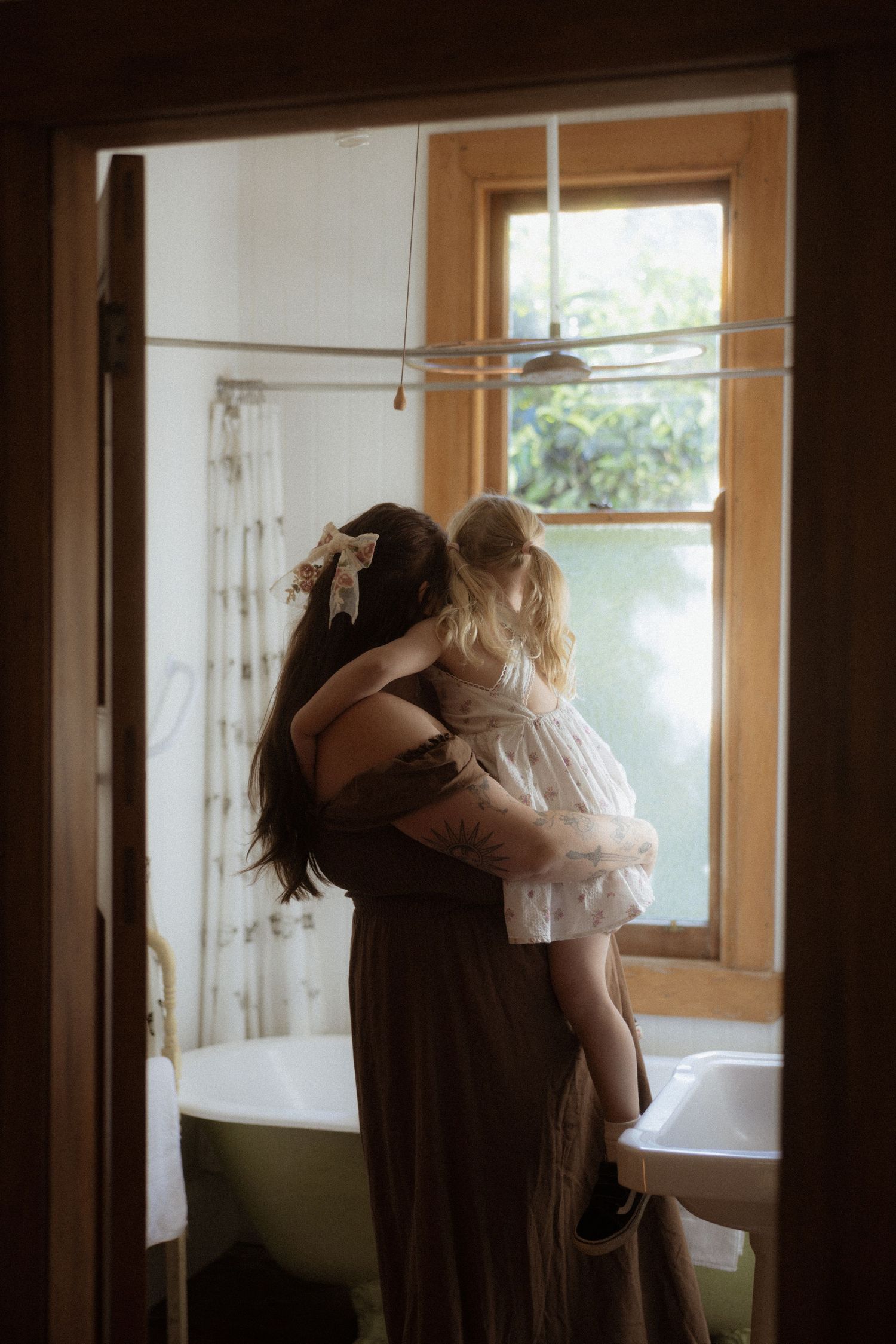 A tender moment captured in a sunlit bathroom with a vintage clawfoot tub and window light filtering through.