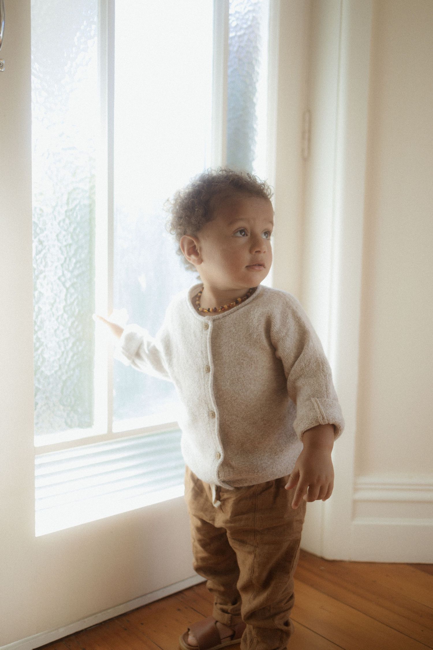 Four sequential images show a small child in a beige sweater and brown pants standing near bright sunlit windows.