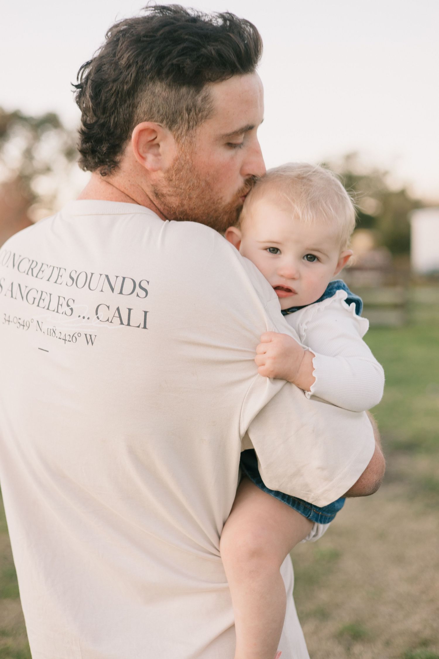 A person wearing a white t-shirt holds and kisses a small child while standing in a field at sunset.