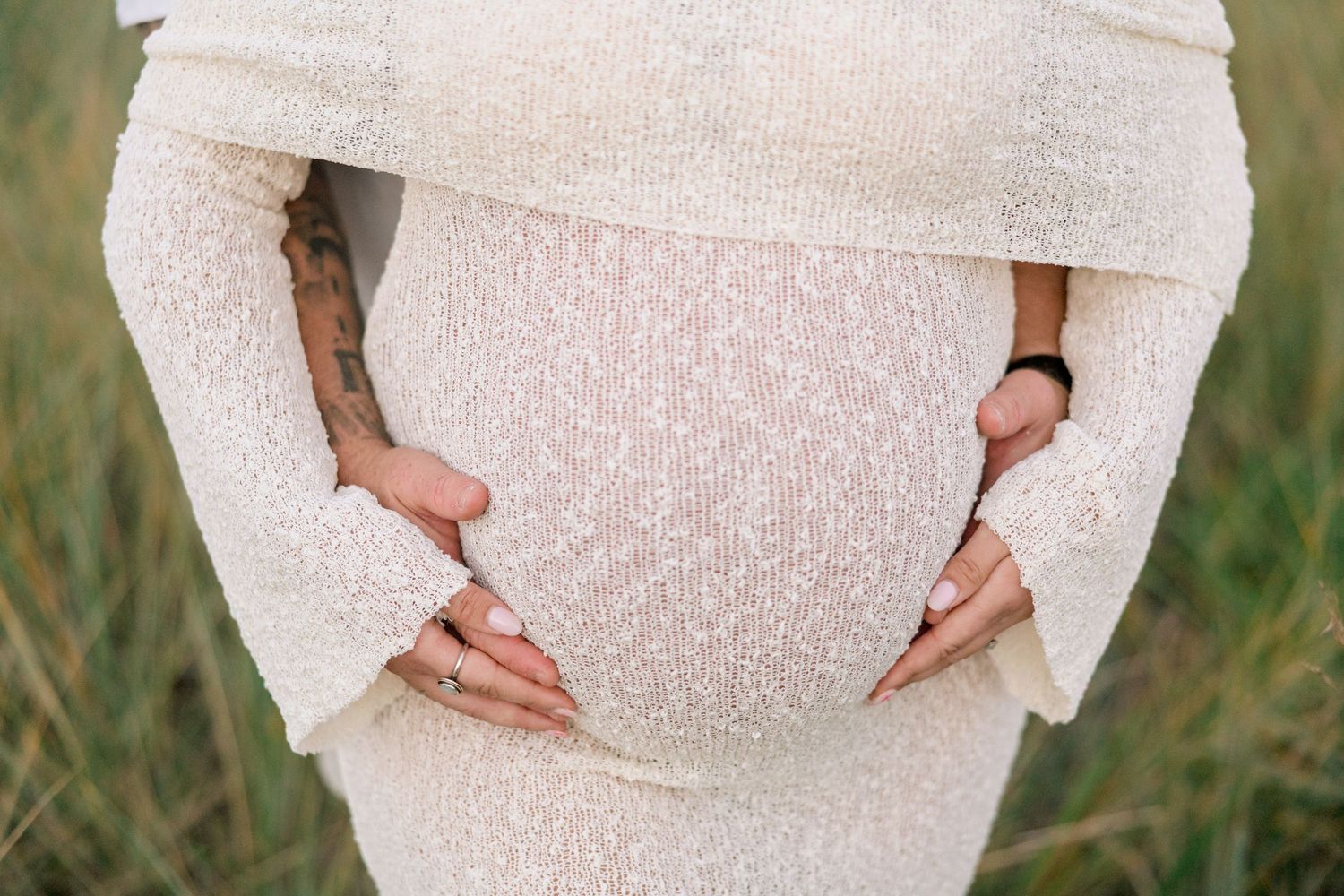 Pregnant woman in white textured dress cradling baby bump outdoors in a green field.
