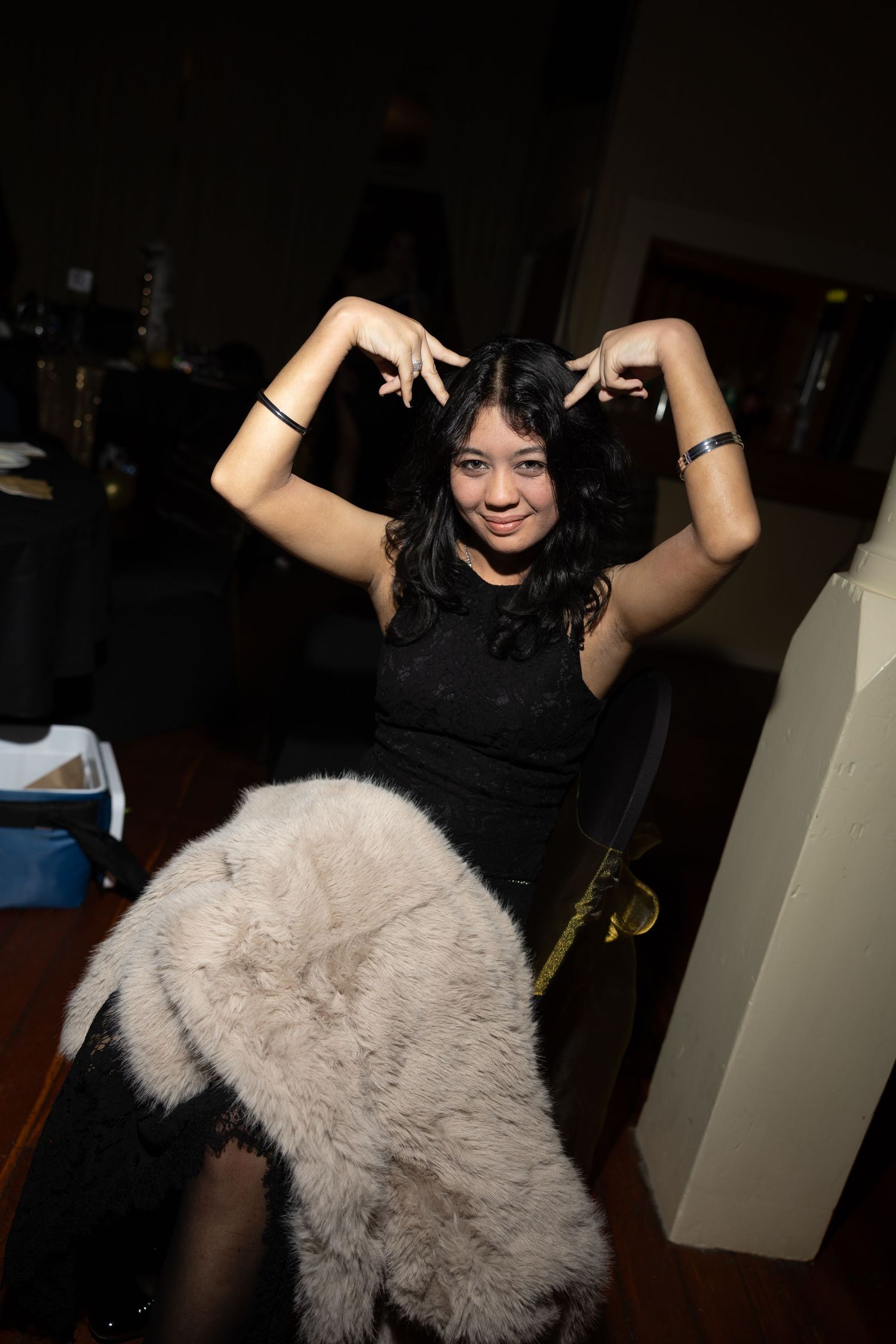 Young woman in black dress posing playfully with hands raised like horns, smiling indoors at a party.