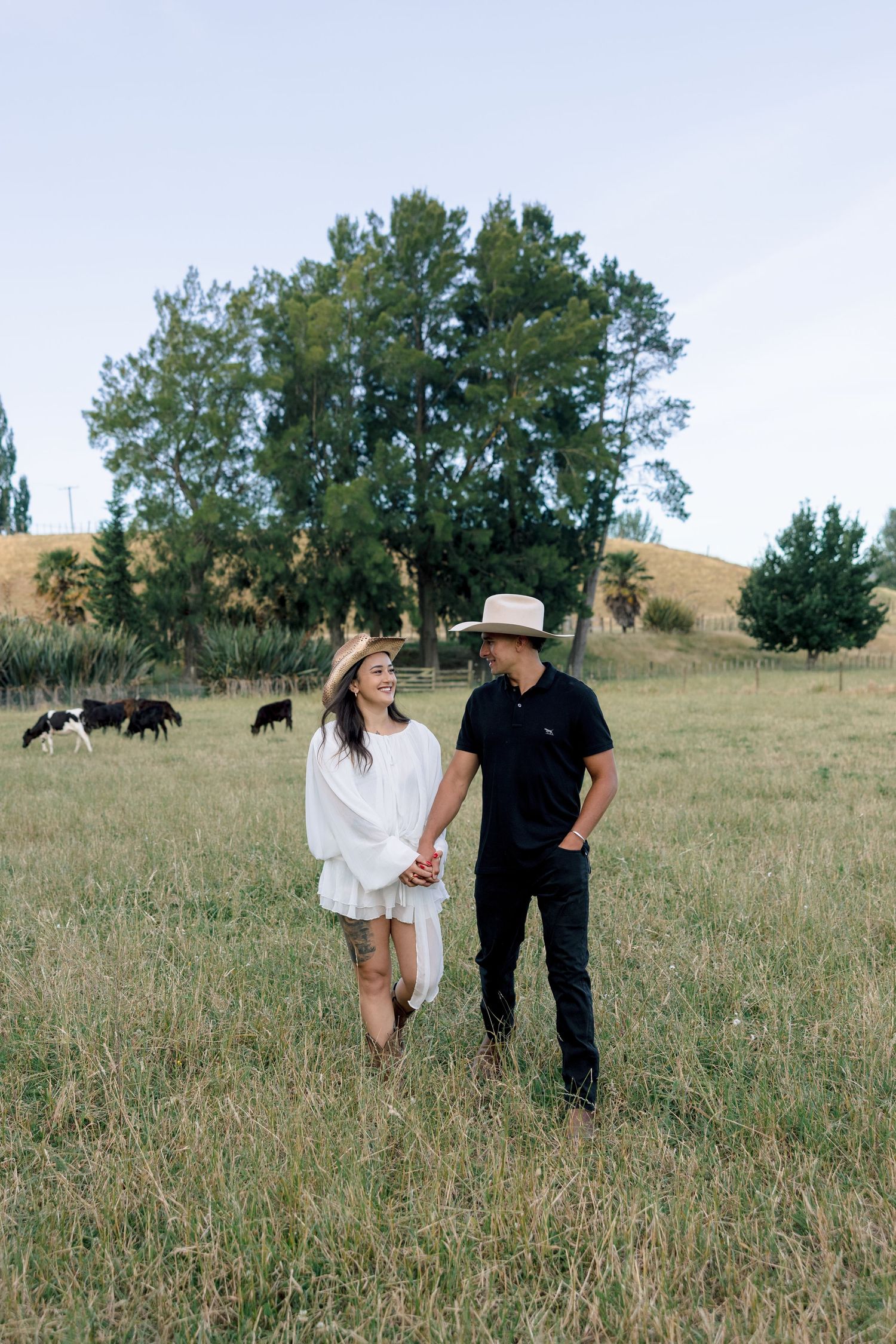 A couple walks hand in hand through a grassy field with grazing cattle and rolling hills in the background.