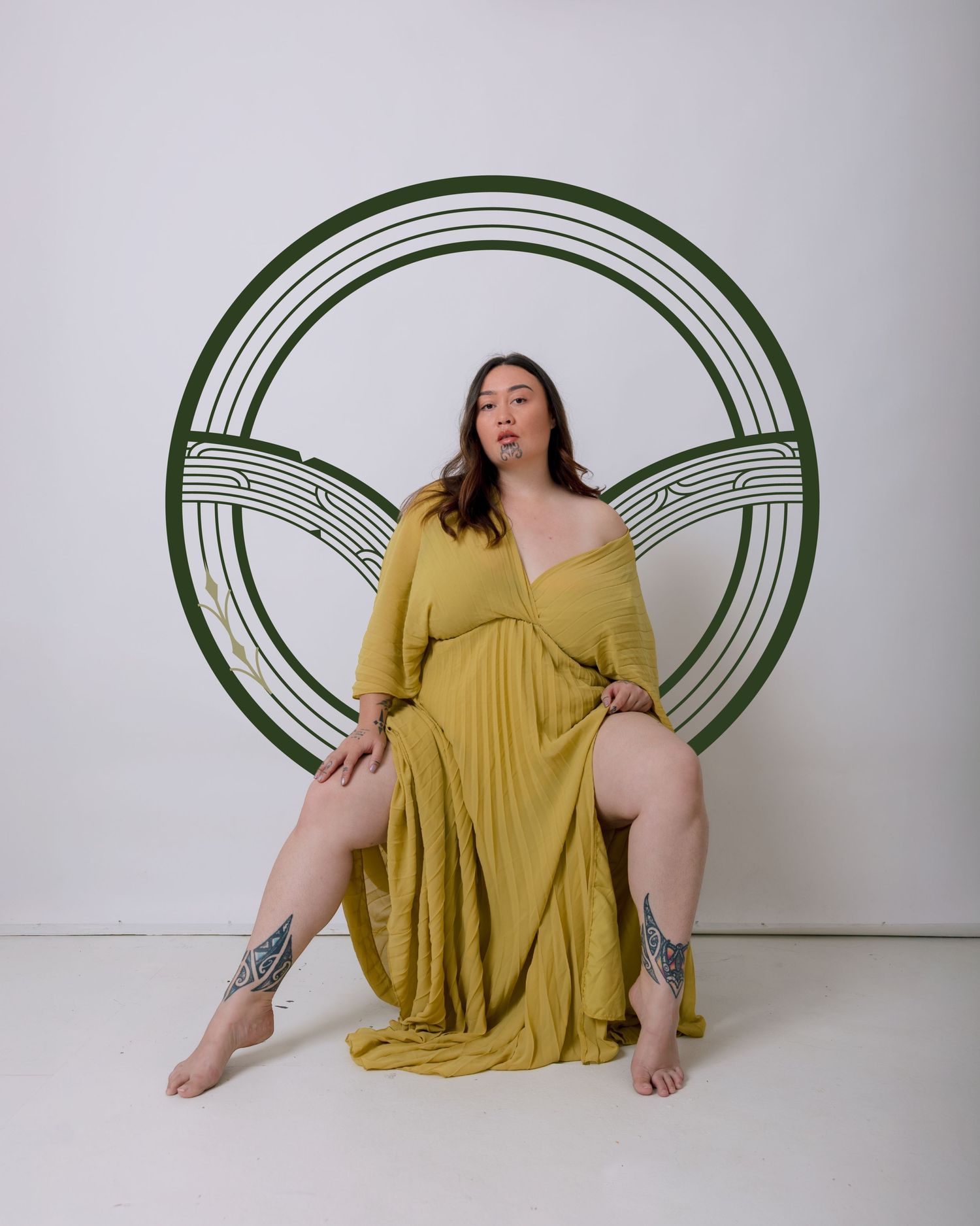 Model in flowing yellow dress poses seated against green circular art backdrop in studio setting.