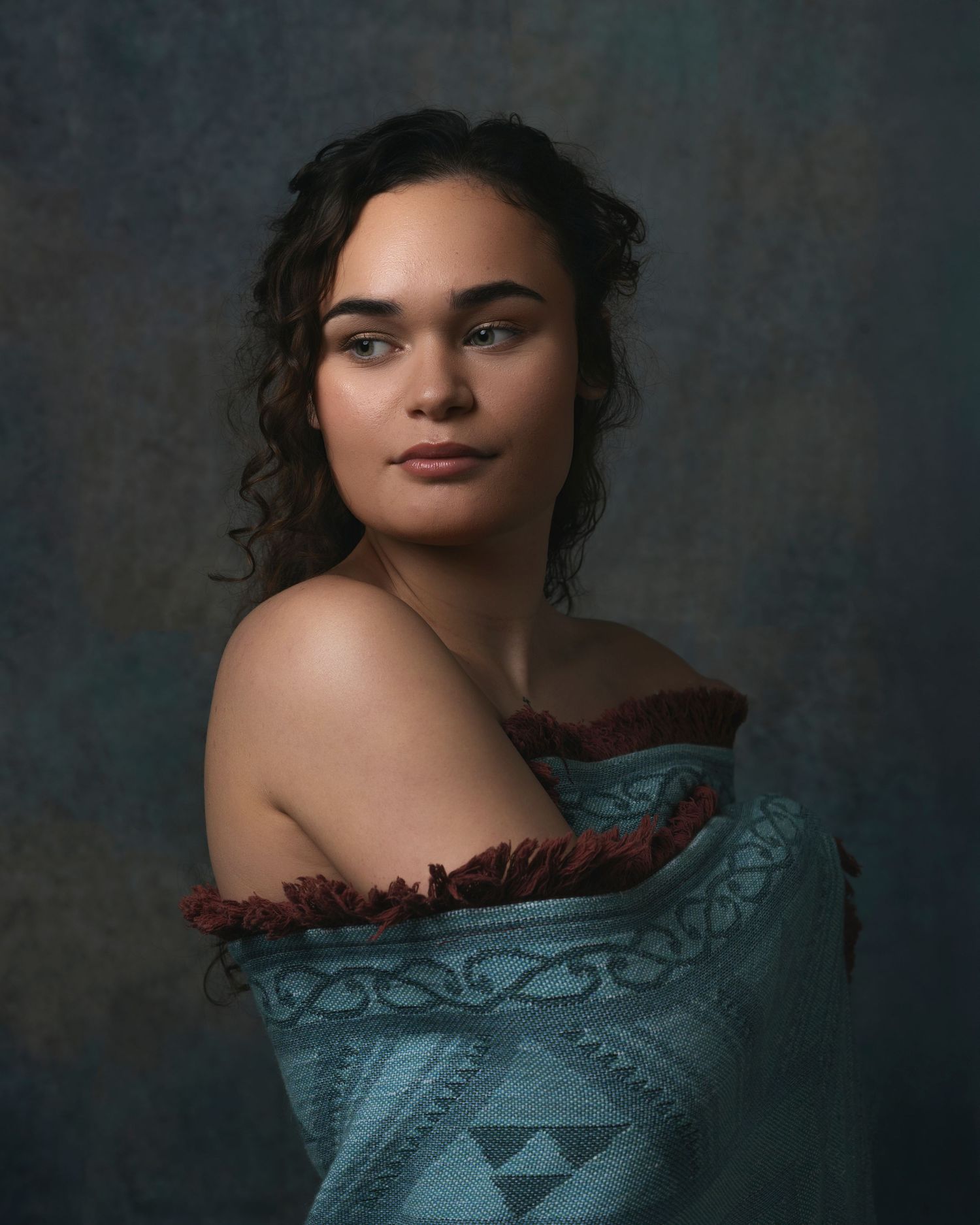 Young woman with curly dark hair draped in a teal embroidered off-shoulder wrap, posed against a moody studio backdrop.