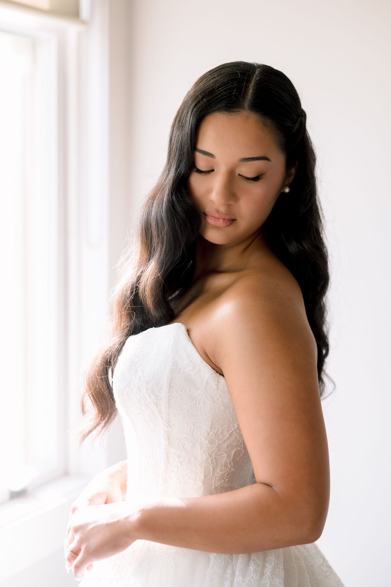 Elegant bride in strapless lace wedding gown with long dark wavy hair, posing gracefully near a bright window.