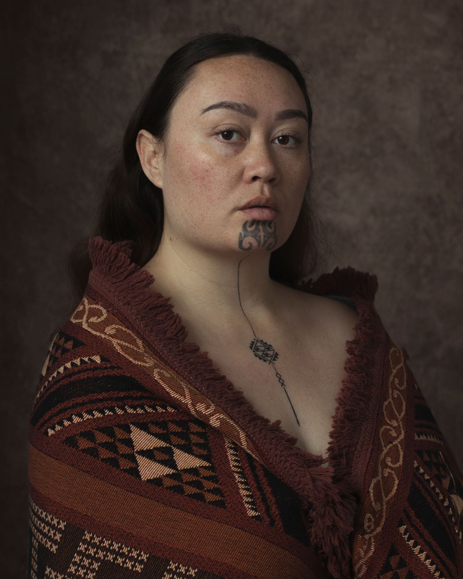 A young Maori woman wearing a traditional korowai cloak with a moko kauae chin tattoo and greenstone pendant.