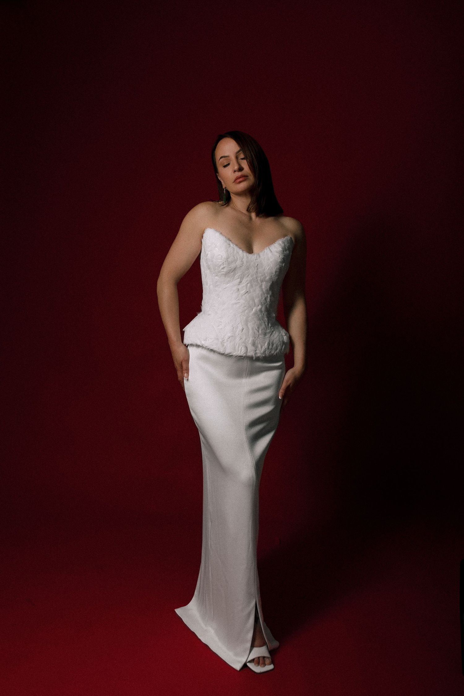 Woman in white lace corset top and satin maxi skirt posing against dark red background.