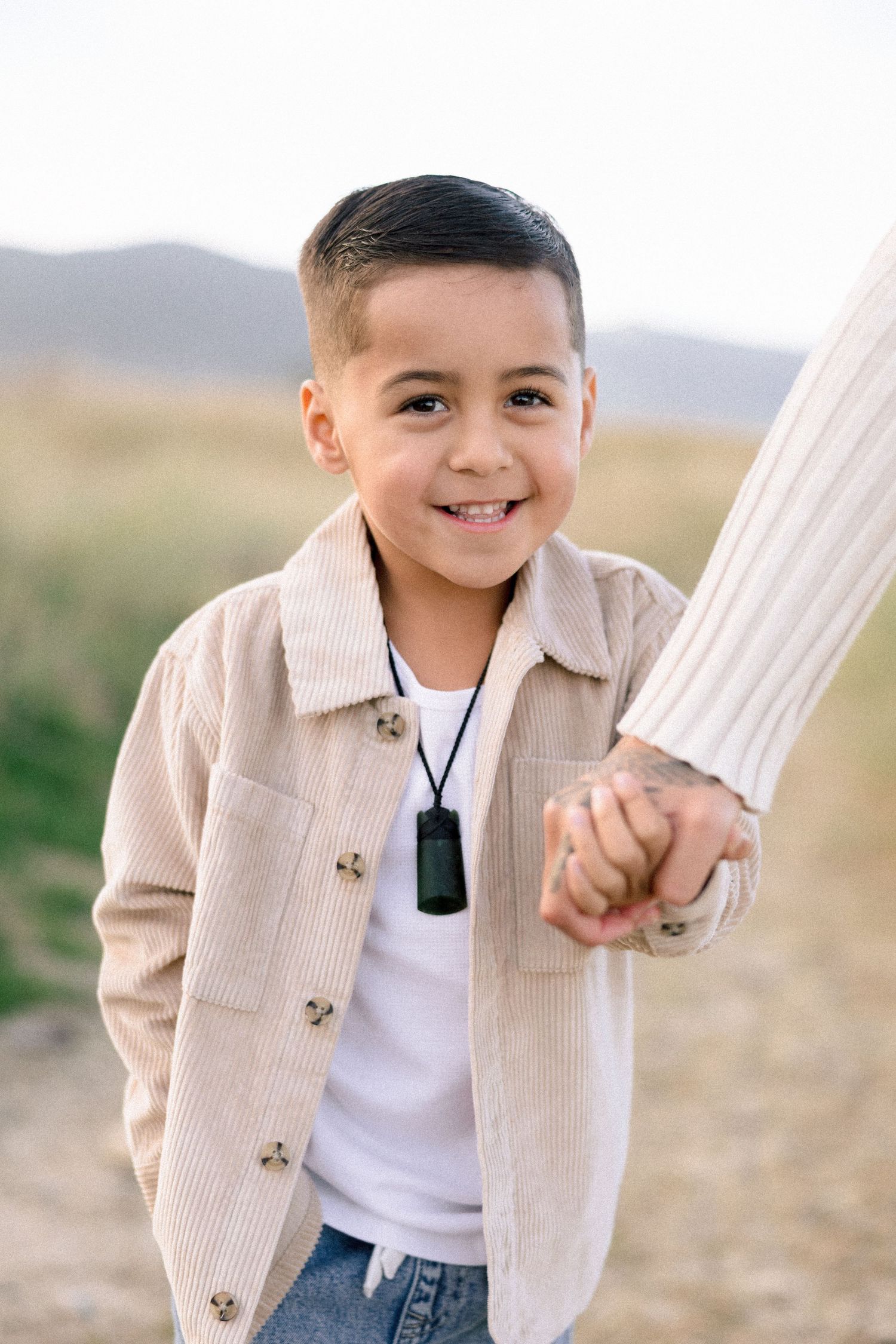 Smiling young boy holding hands with adult outdoors, wearing beige corduroy jacket and dog tag necklace on scenic path.