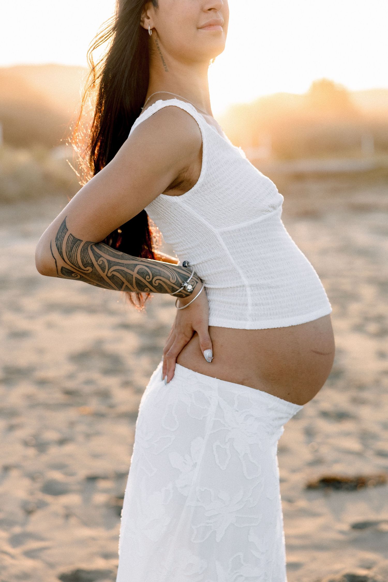 Pregnant woman in white dress and crop top with tattoo on arm poses on sandy beach at golden sunset.