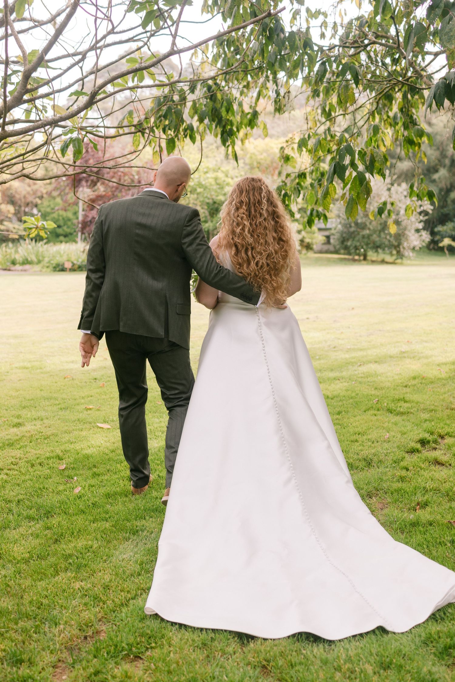 A couple in wedding attire walk together on a lush green lawn beneath overhanging tree branches.