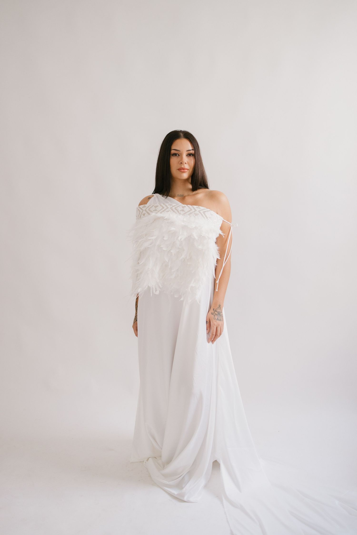 Model wearing an elegant white one-shoulder wedding gown with lace details and flowing train against light background.