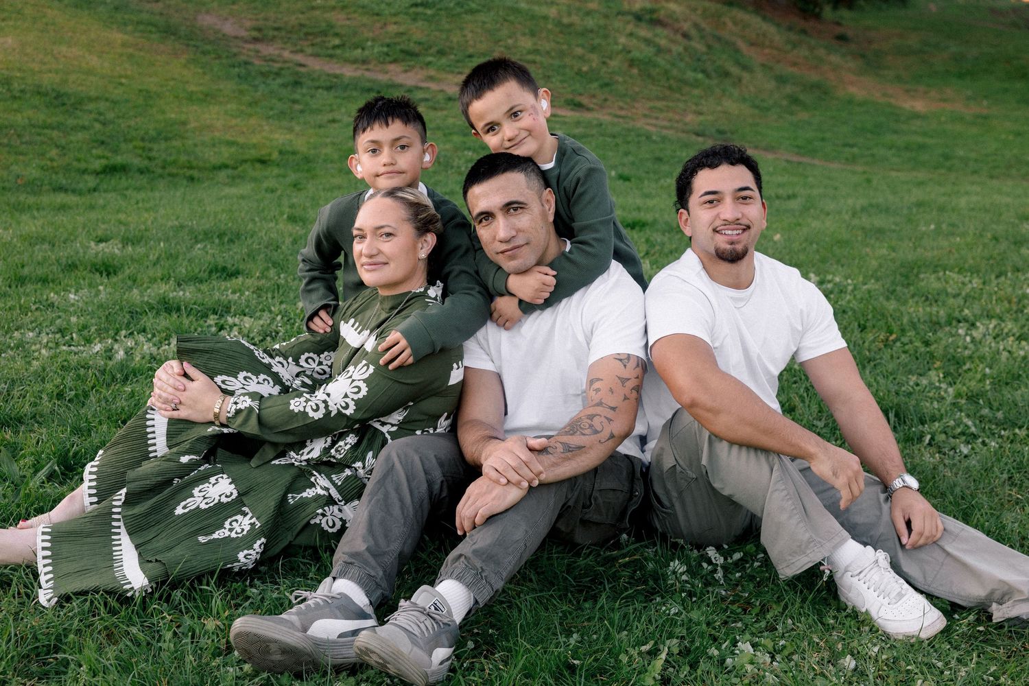 Family of five smiling together on green grass, with two boys sitting behind their parents during an outdoor photo session.