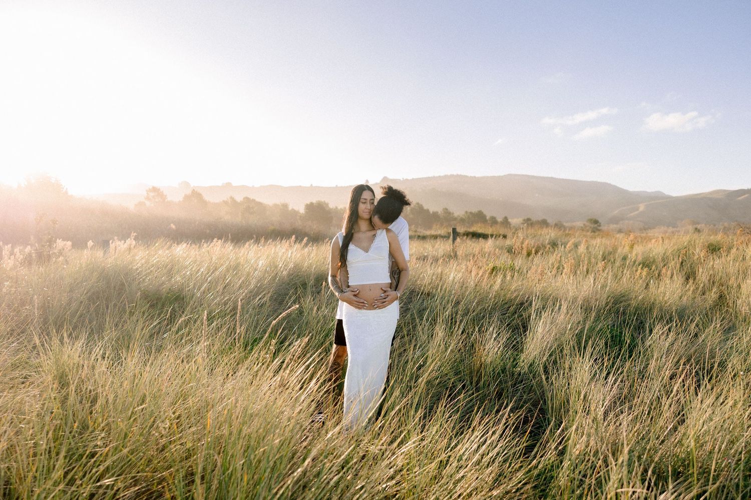 Pregnant couple embracing in golden grass field with mountain backdrop at sunset, woman in white dress.