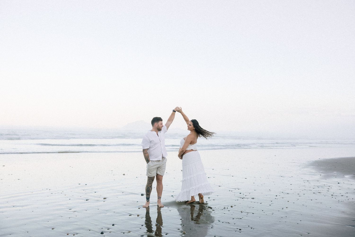 A couple in white outfits dancing on a beach at sunset, holding hands raised, with ocean waves in the background.