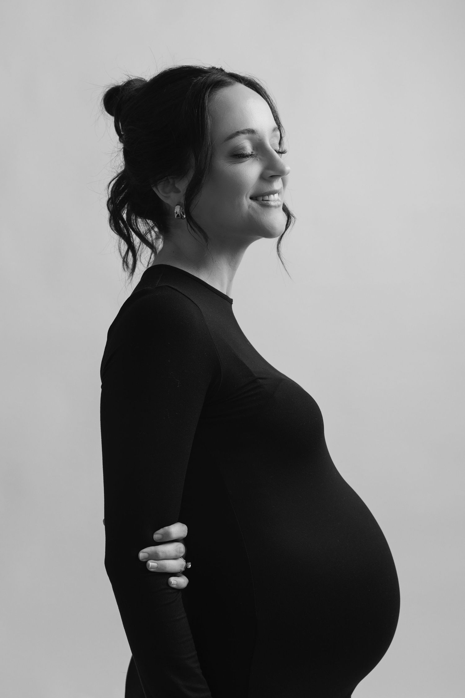 Black and white portrait of a smiling pregnant woman in a black dress, cradling her baby bump with eyes closed.