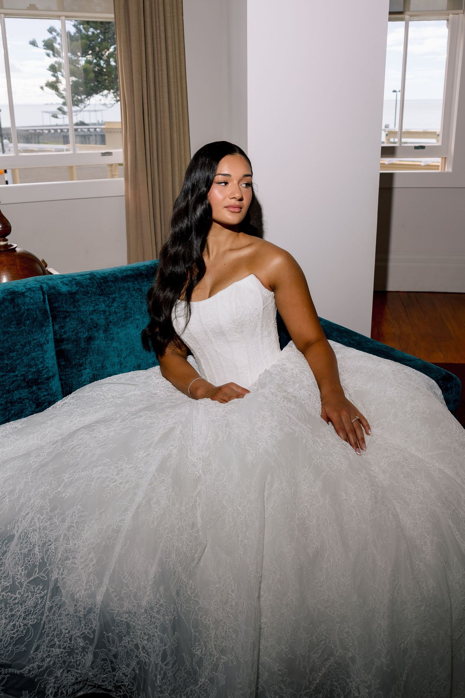 A bride in a stunning strapless white ballgown with lace details sits gracefully on a teal velvet sofa.