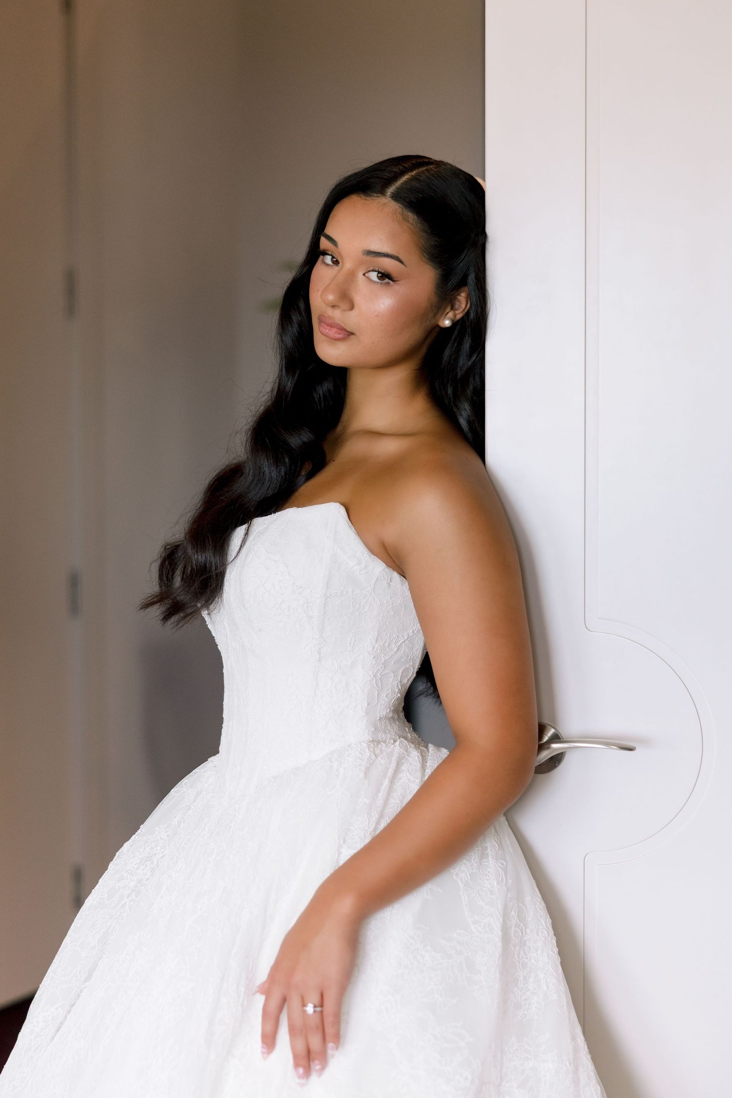 Bride in strapless white ballgown wedding dress leaning against white door with long dark wavy hair.