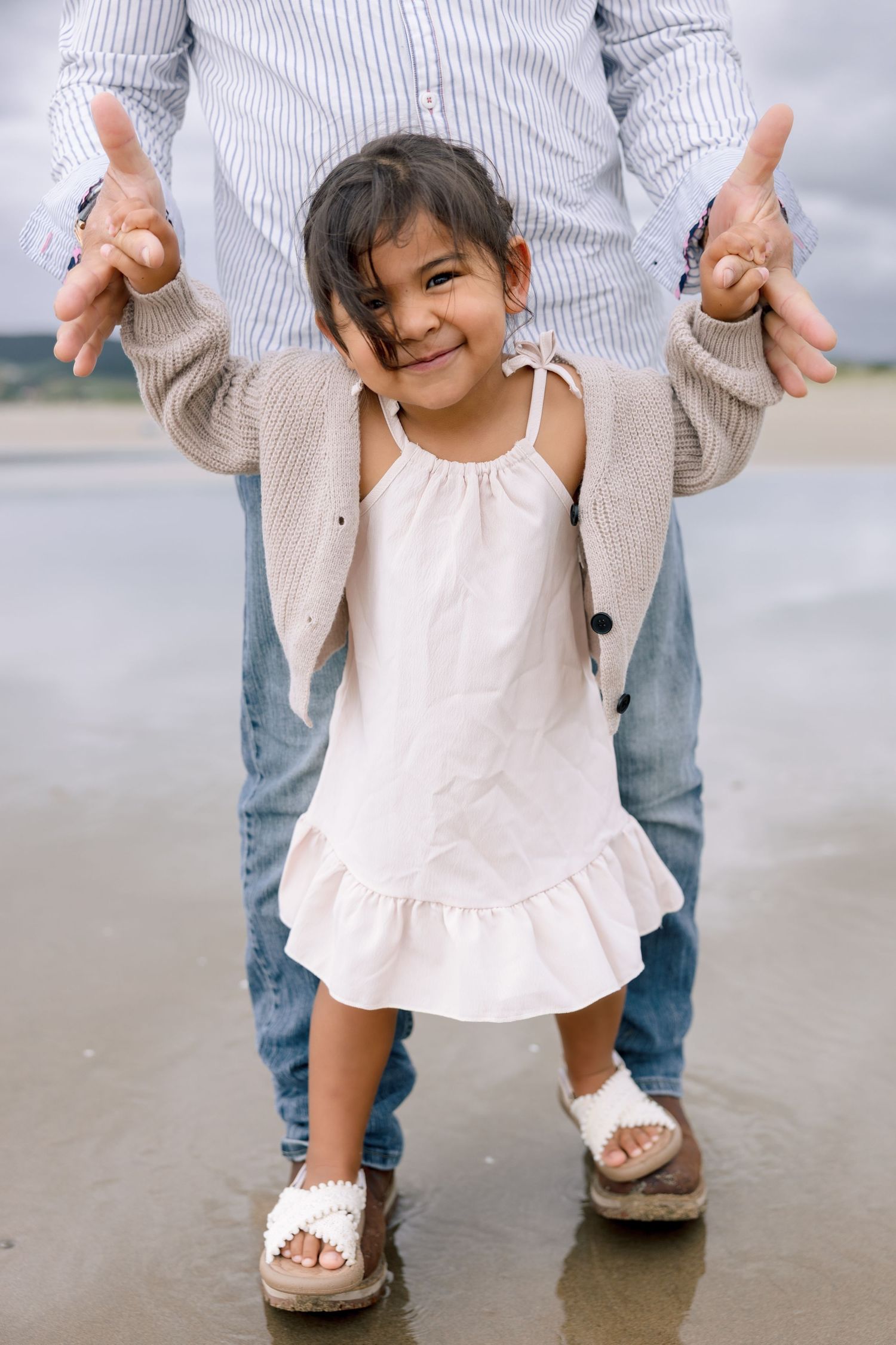 A child in a white dress holding hands with an adult wearing jeans and a striped shirt on a light colored floor.