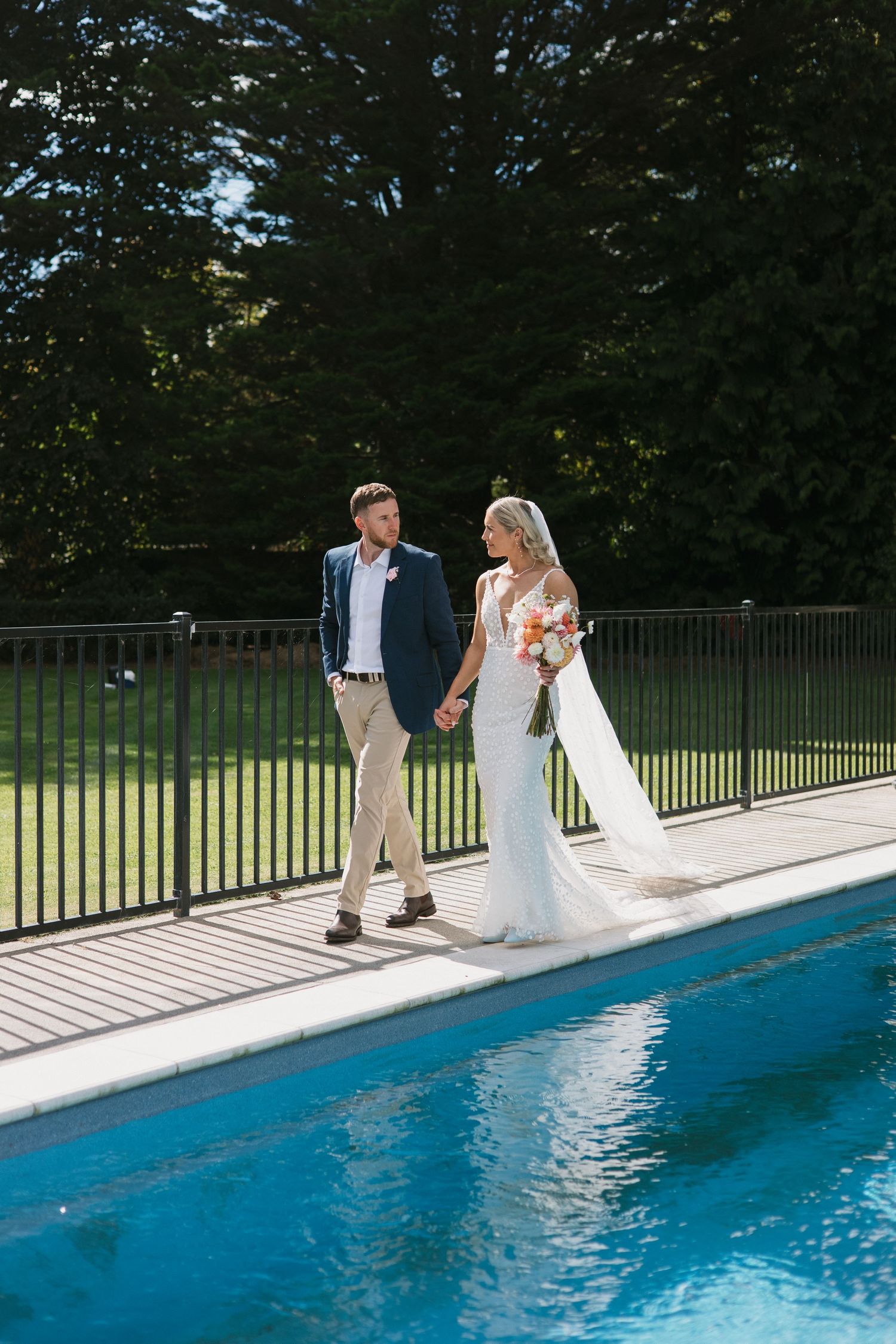 A couple in wedding attire shares a romantic moment while standing beside a bright blue swimming pool on a sunny day.