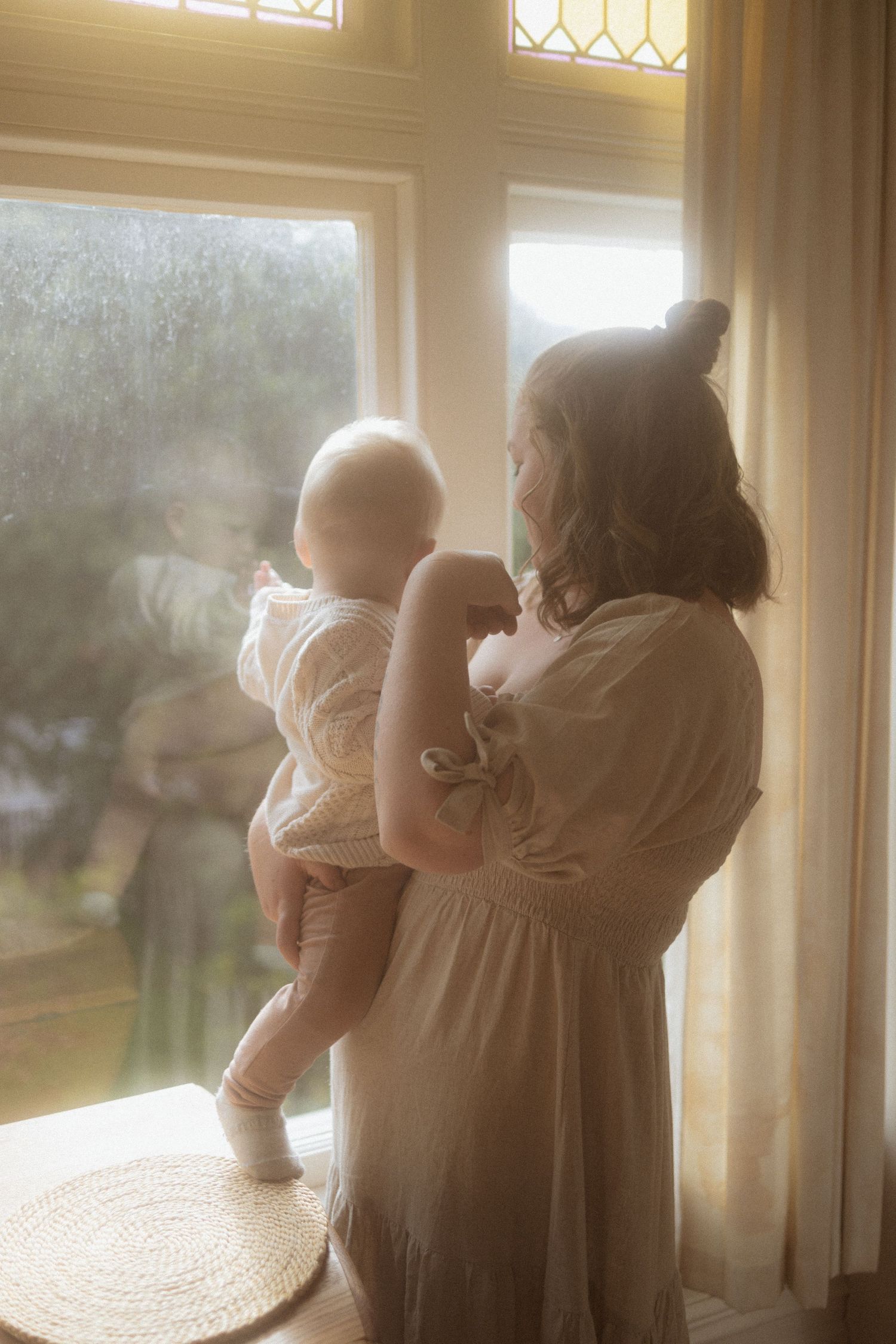 A silhouetted figure in a flowing dress holds a baby while standing by a sunlit window in a dreamy, atmospheric scene.