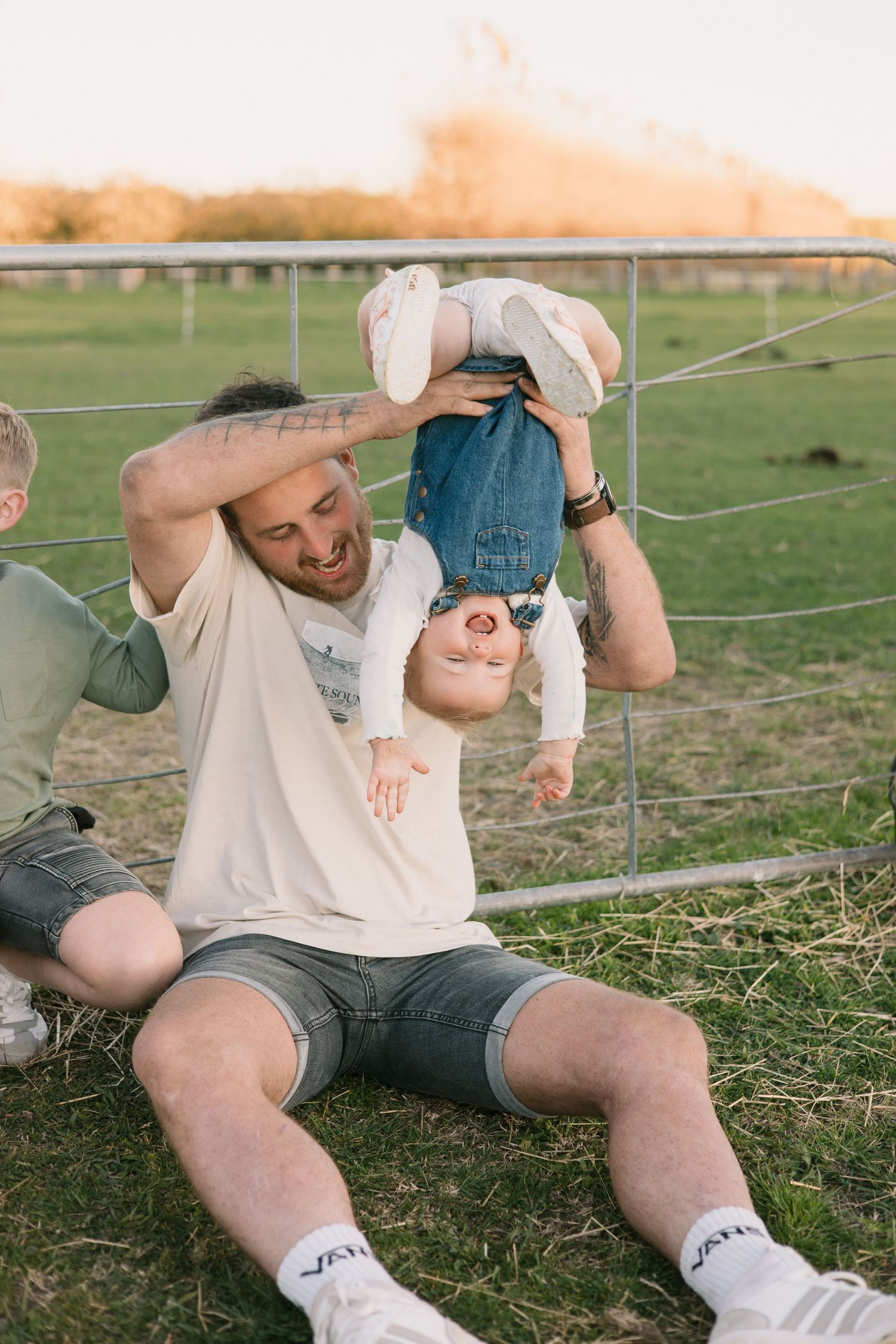 A playful moment captured outdoors as a family member holds a laughing child upside down by a wire fence at sunset.