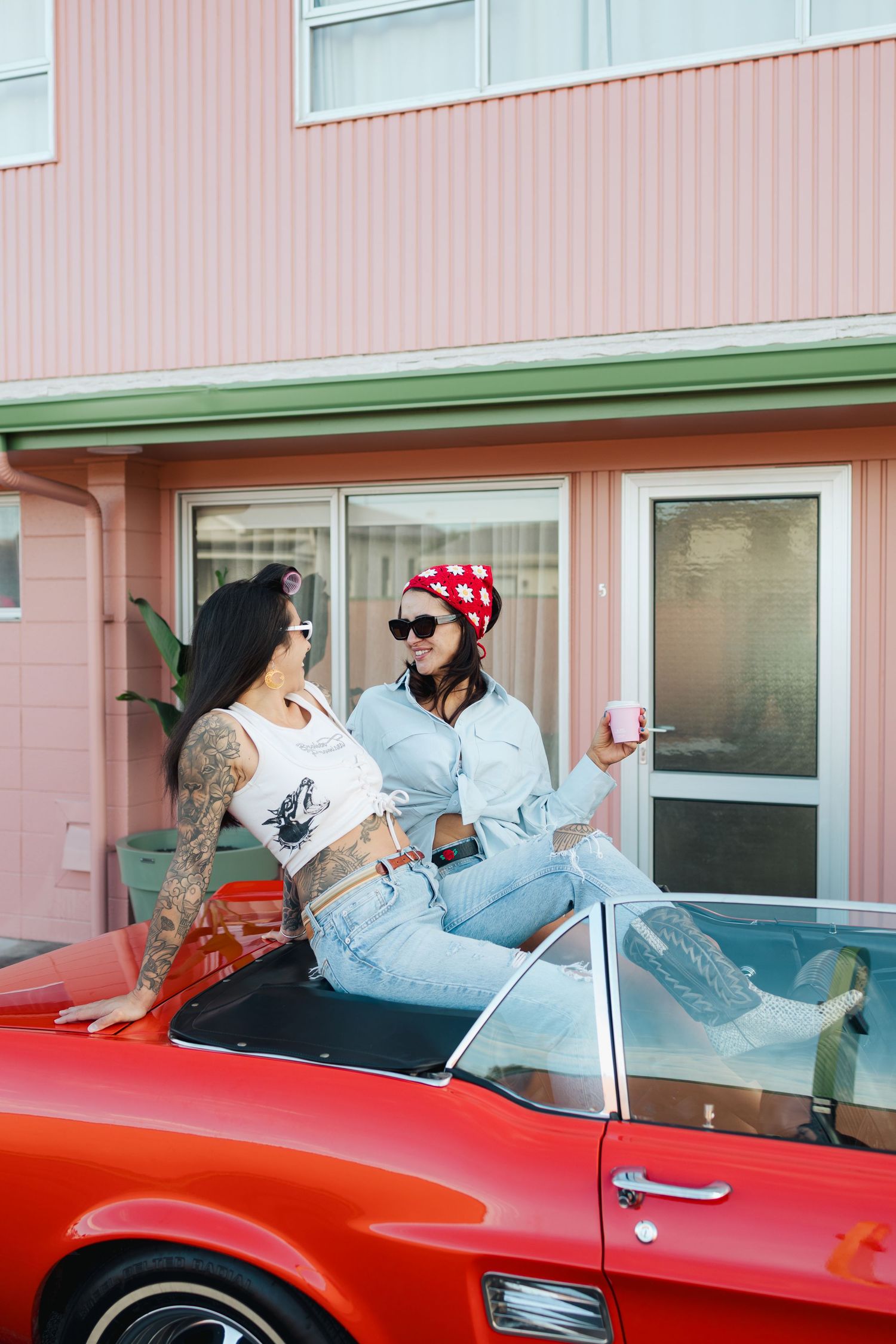 Two friends sitting on vintage red car having drinks outside pink motel.
