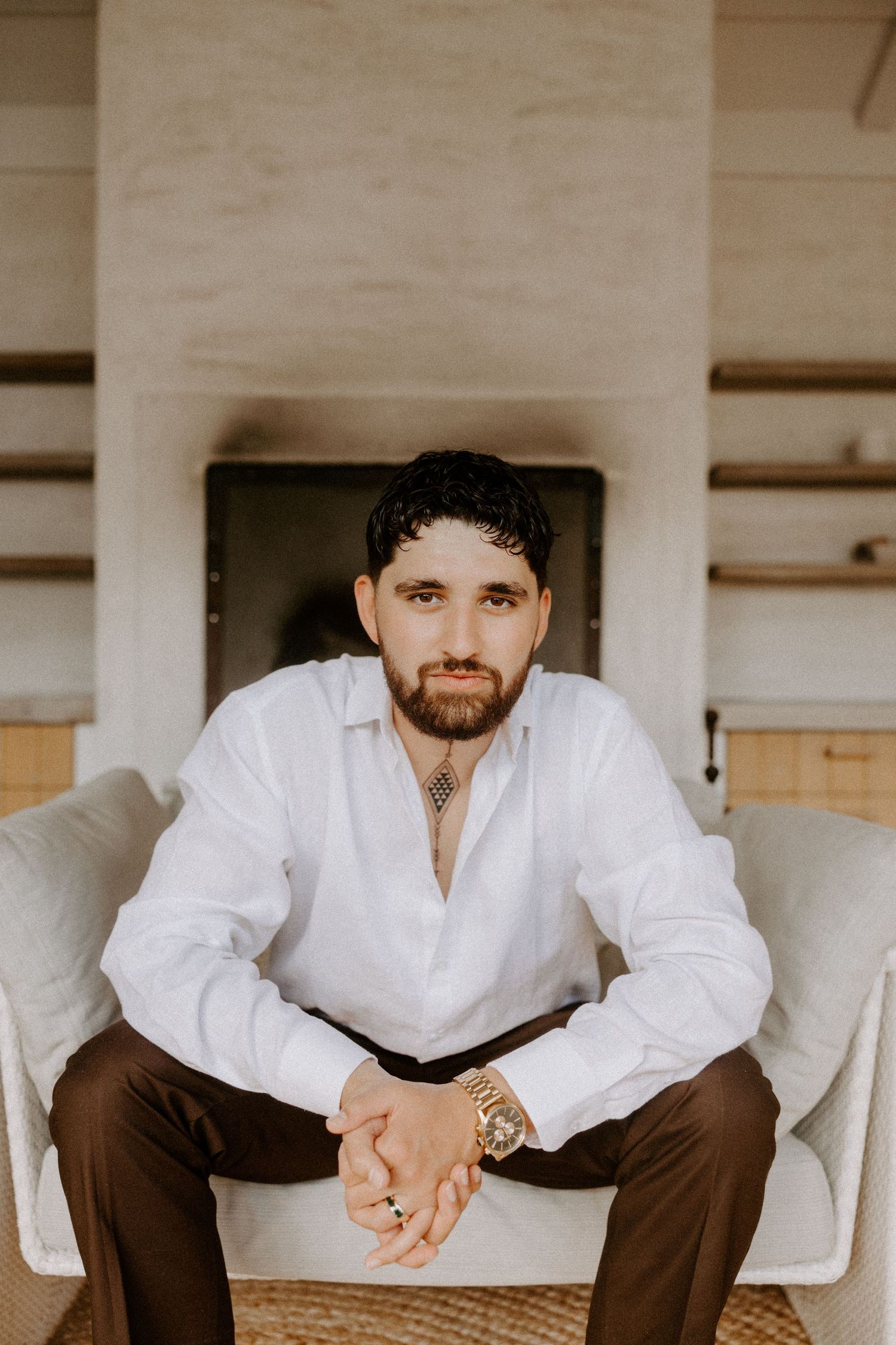 A portrait of someone in a white button-down shirt and brown pants sitting in a light-colored chair against a beige wall.