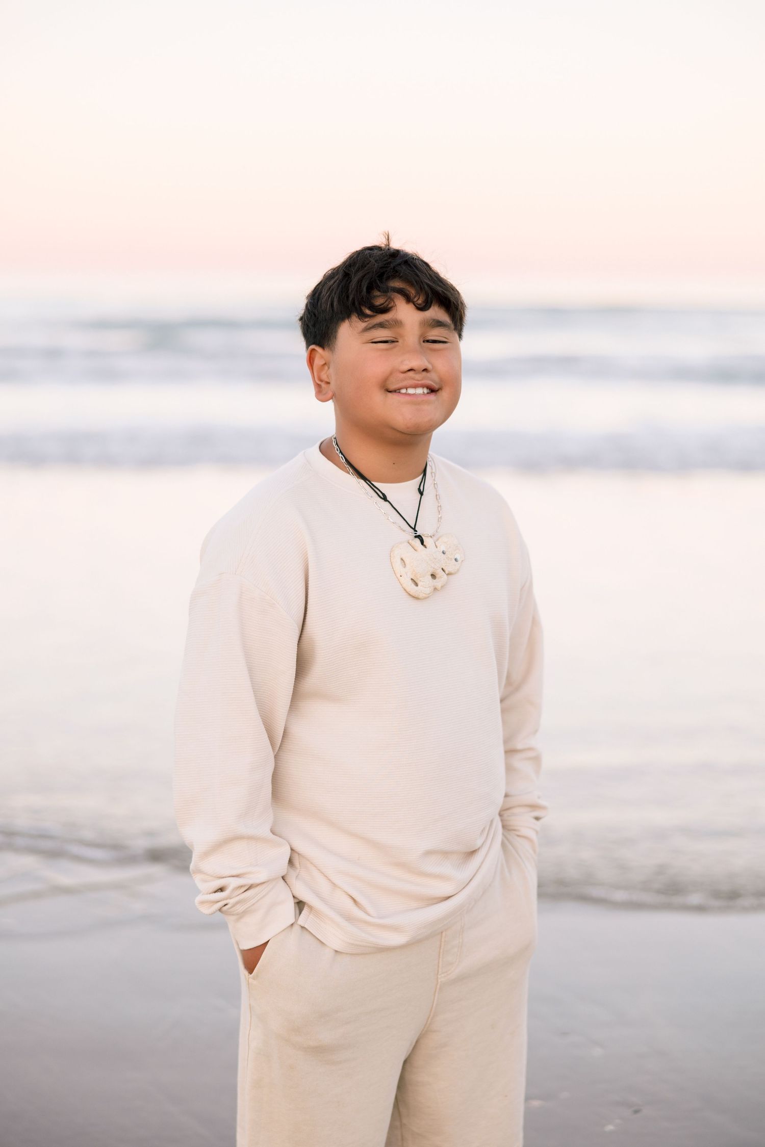 Smiling Polynesian boy in beige outfit wearing a bone carving necklace stands on a beach at sunset.