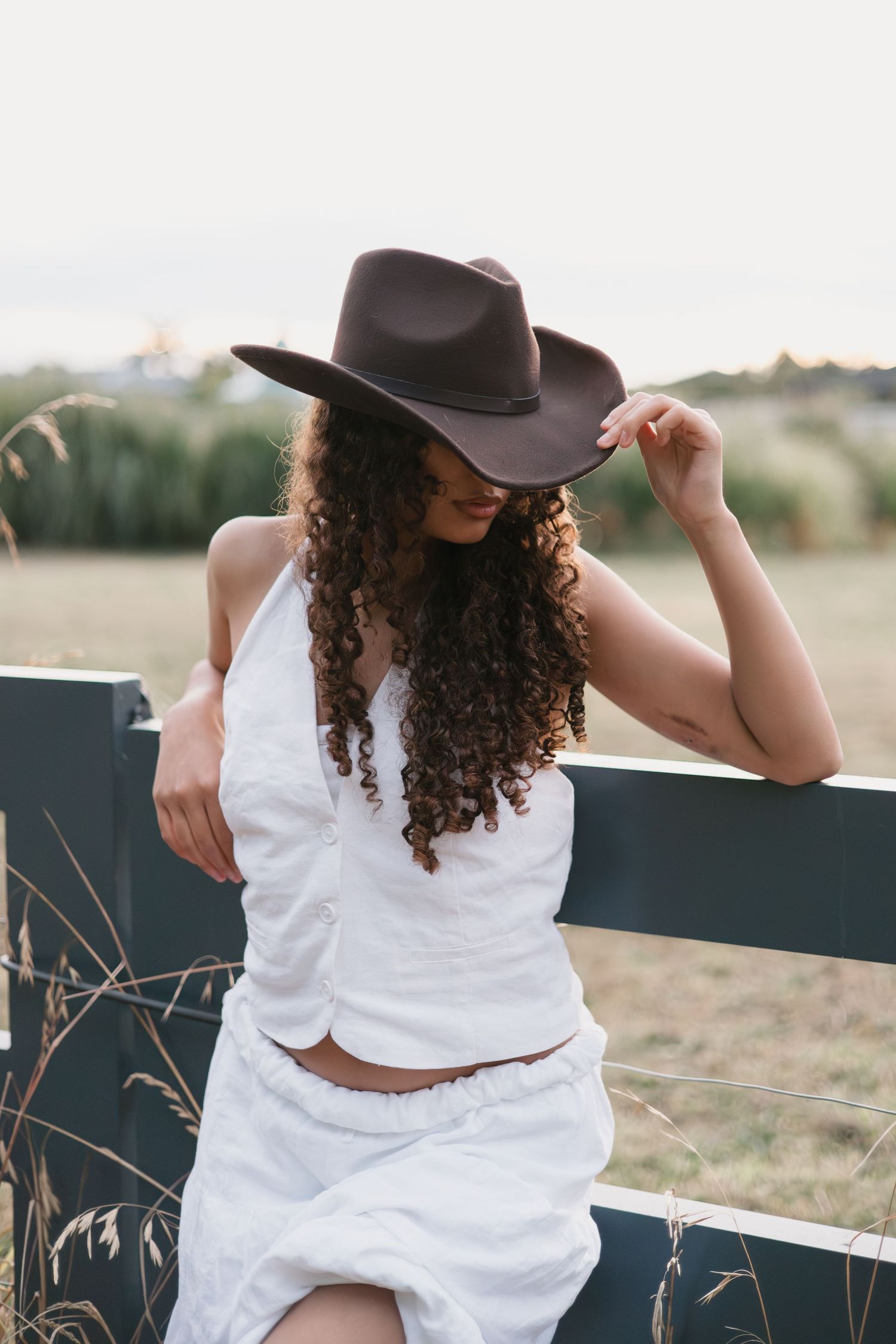Person in white dress and brown hat poses against rustic fence at sunset in countryside setting.