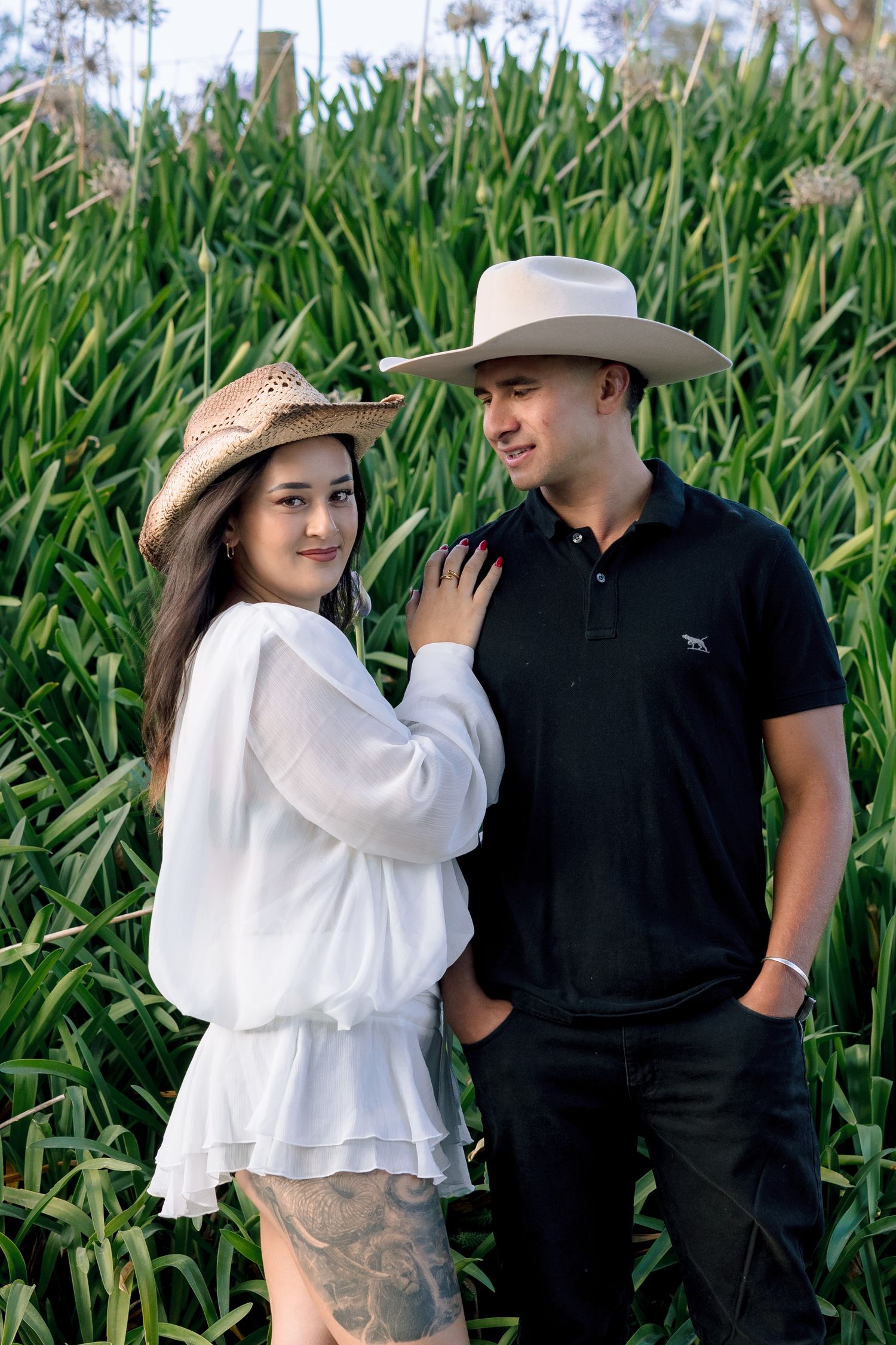 A couple poses together in front of tall green corn stalks, with the woman wearing a white dress and sun hat.