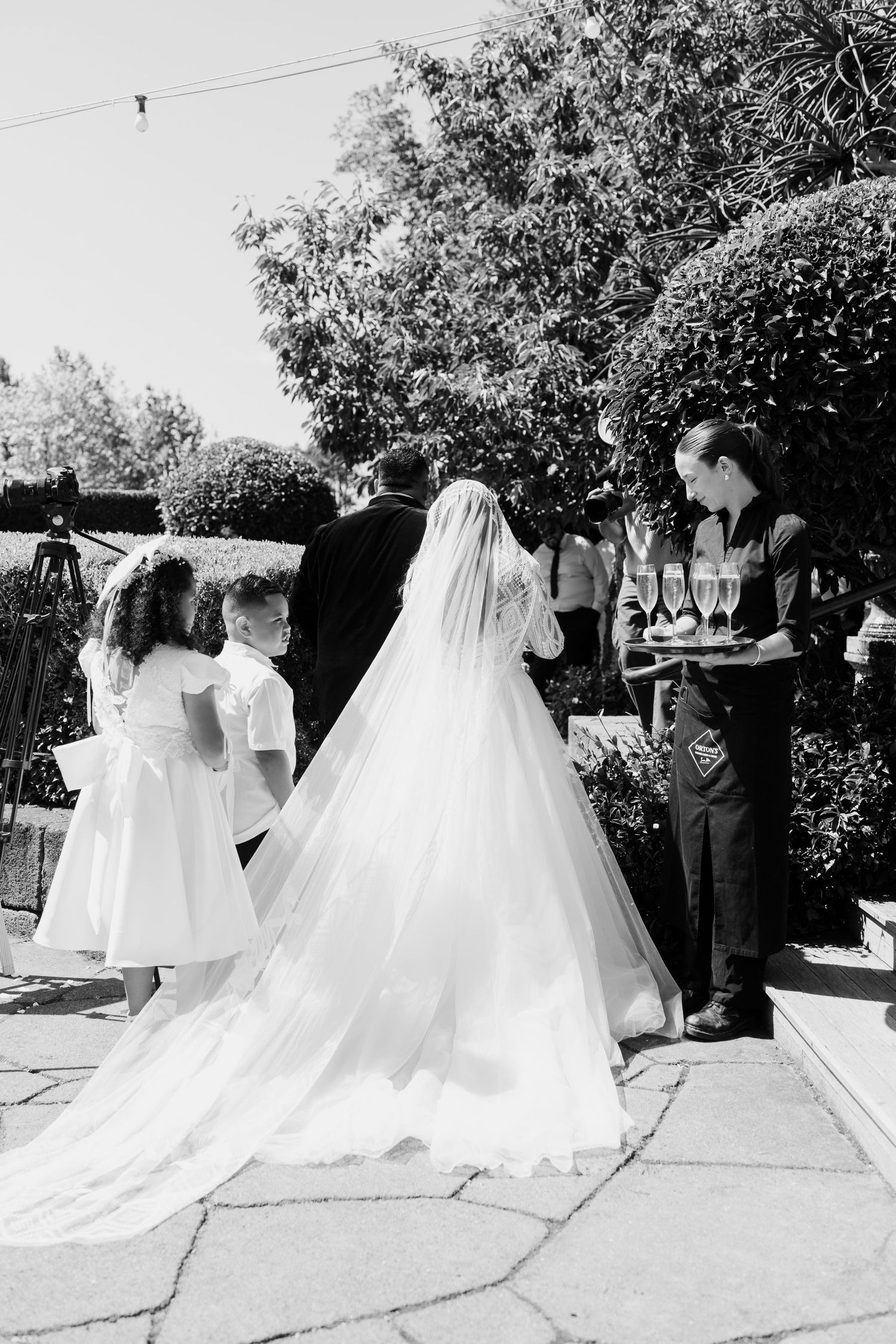 A black and white wedding ceremony photo showing a bride in a flowing white gown with a long train outdoors.