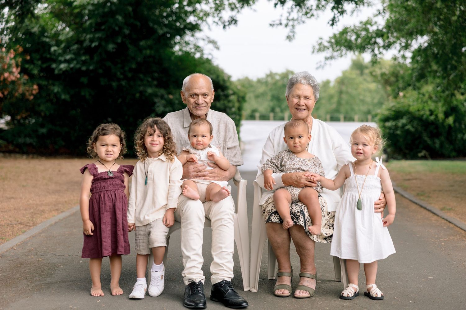 Children sit together on a wooden bench in a park wearing coordinated white and beige outfits.