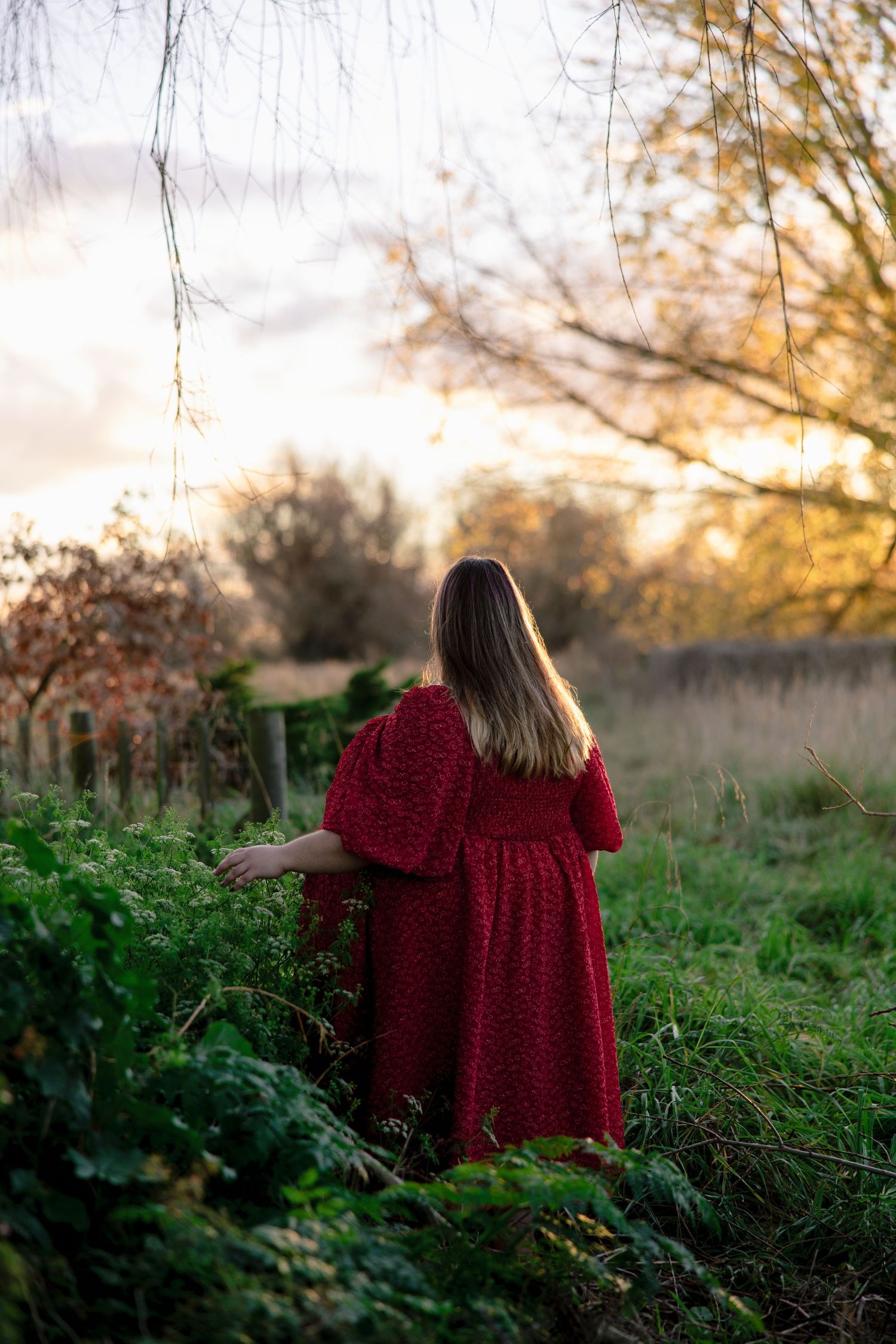 Woman in red dress standing in lush green countryside at golden sunset.
