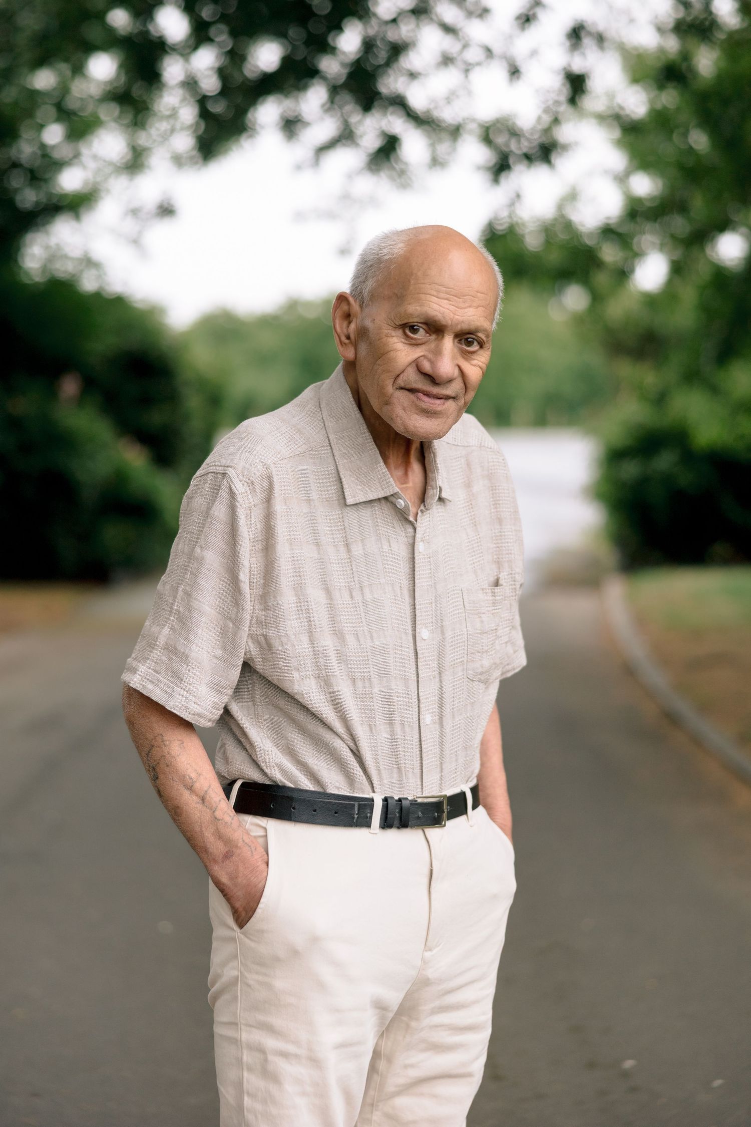 Elderly person wearing light colored clothing stands outdoors on a path surrounded by greenery in a casual pose.