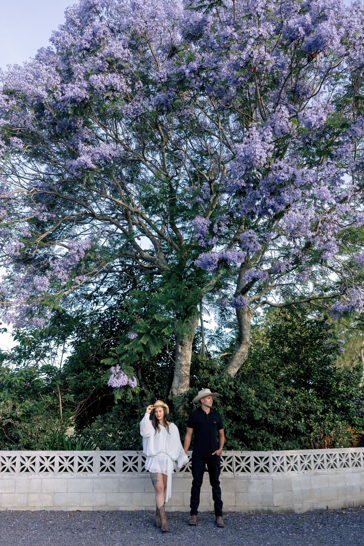 A couple stands beneath a blooming purple jacaranda tree next to an ornate white wall on a sunny day.