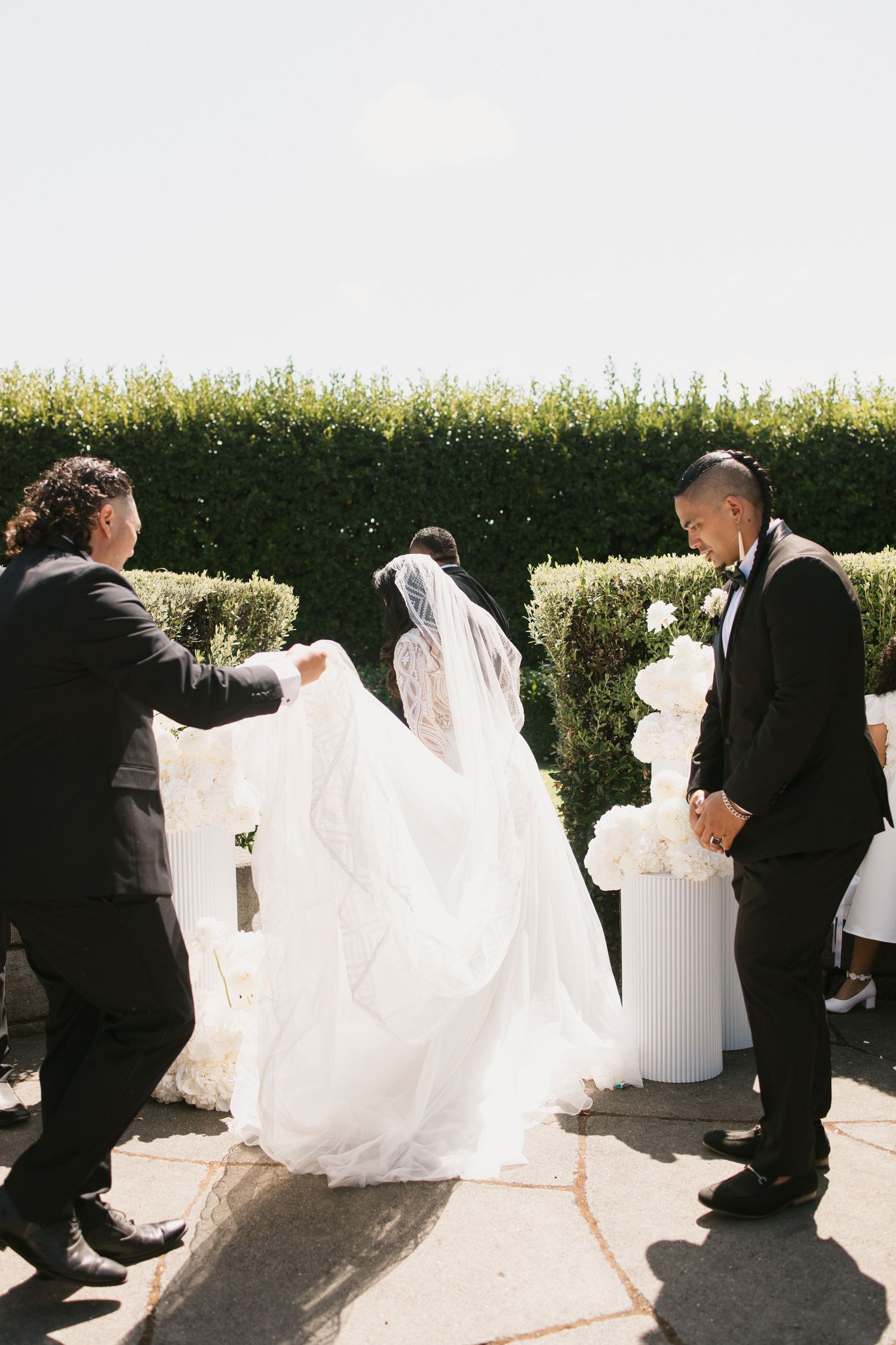 A wedding party helps carry a bride's flowing white dress train across a sunny outdoor patio space.