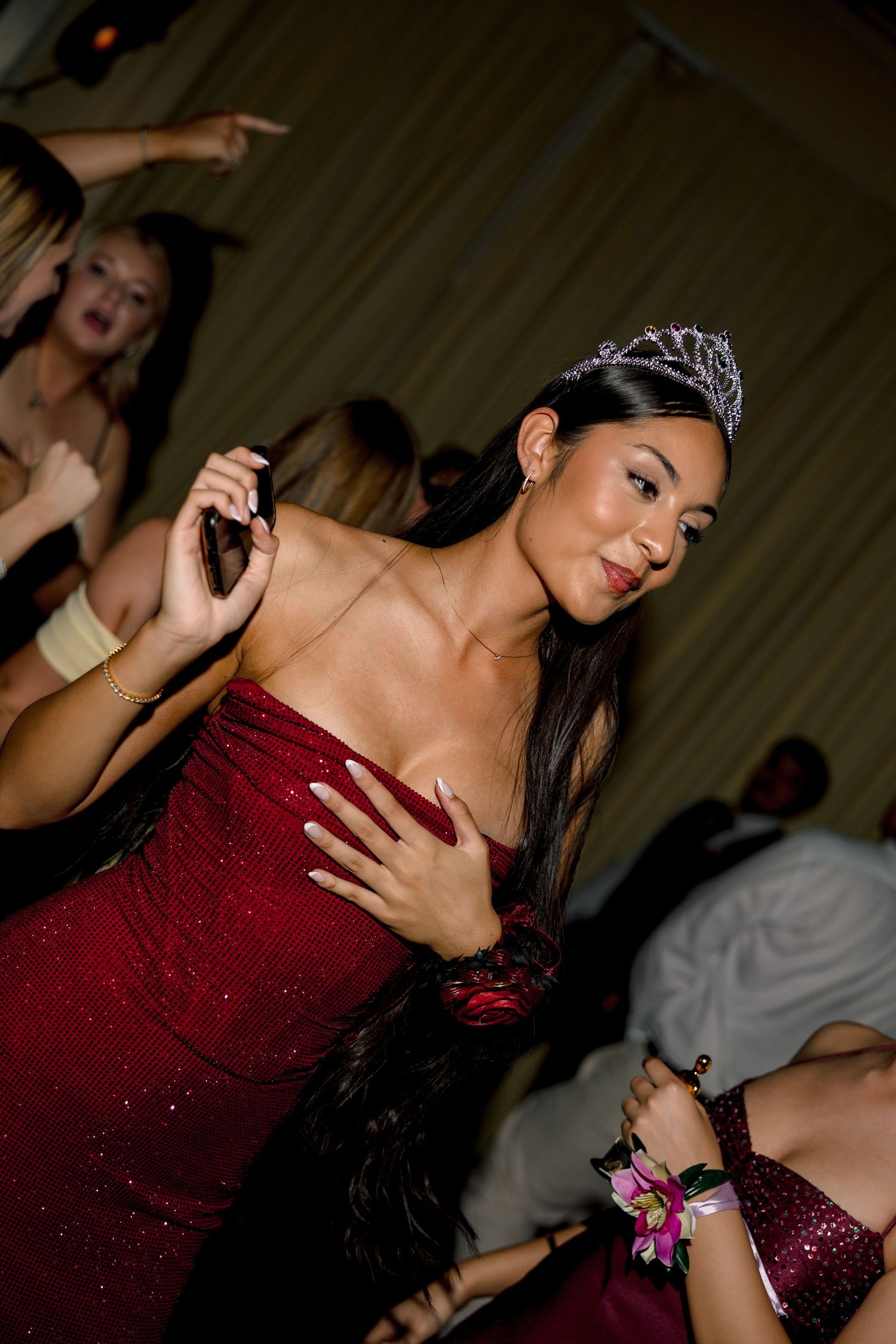 Young woman in red gown wearing tiara dances at formal event surrounded by guests.