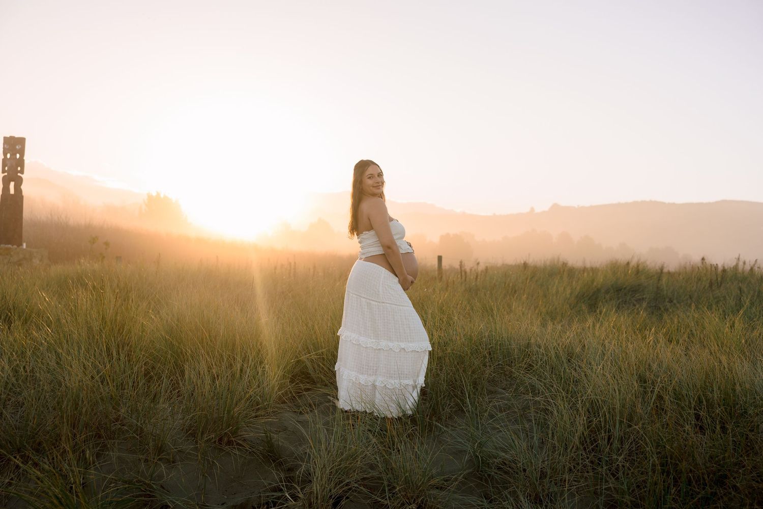 Pregnant woman in white dress standing in golden sunset light among tall grass with mountain backdrop.