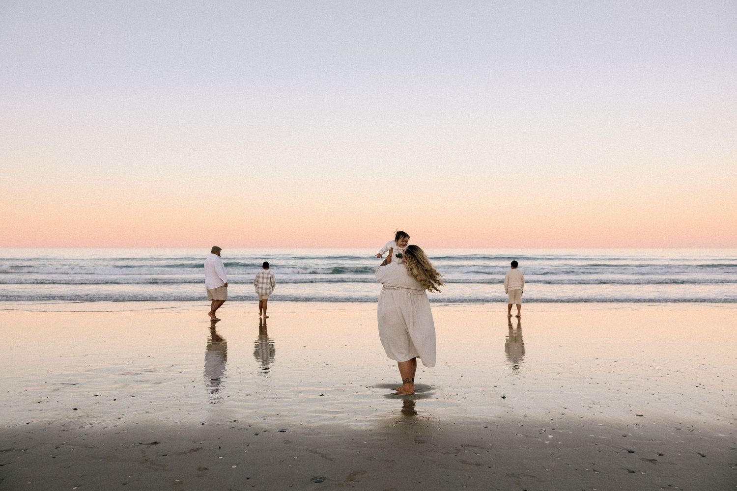 Family in white outfits walking on a reflective beach at sunset with pastel sky and ocean waves.