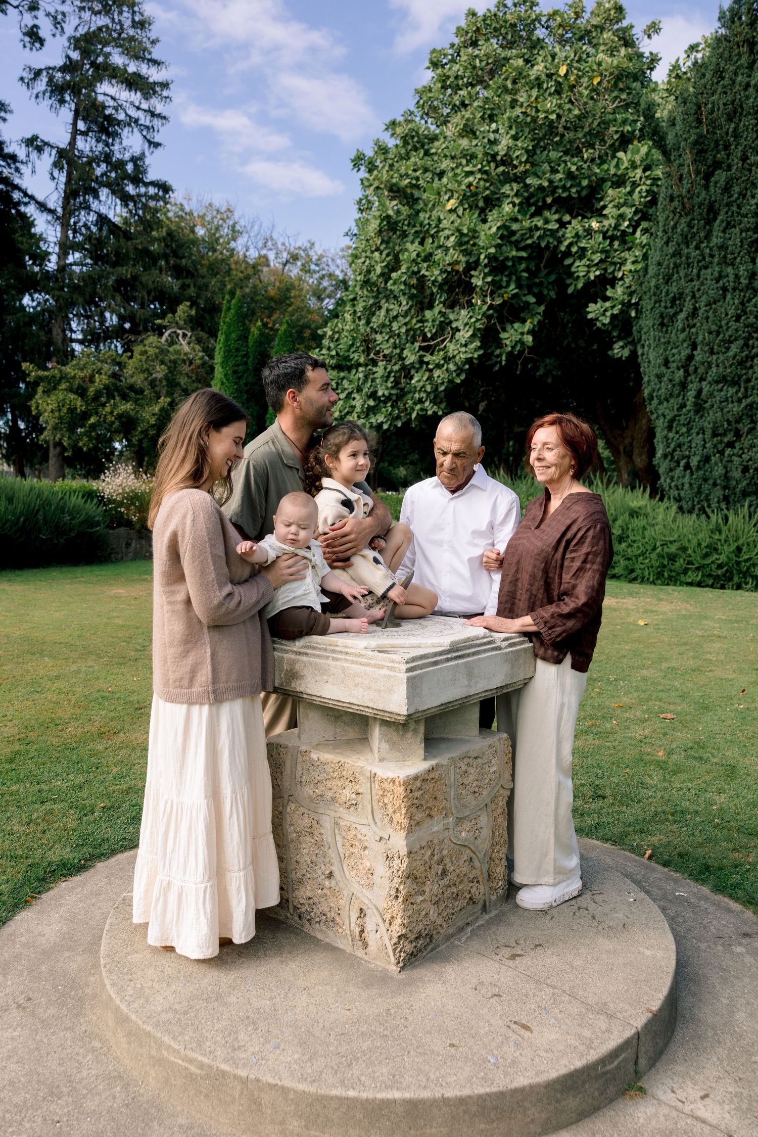 Multi-generational family gathered around a stone pedestal in a lush green park on a sunny day.