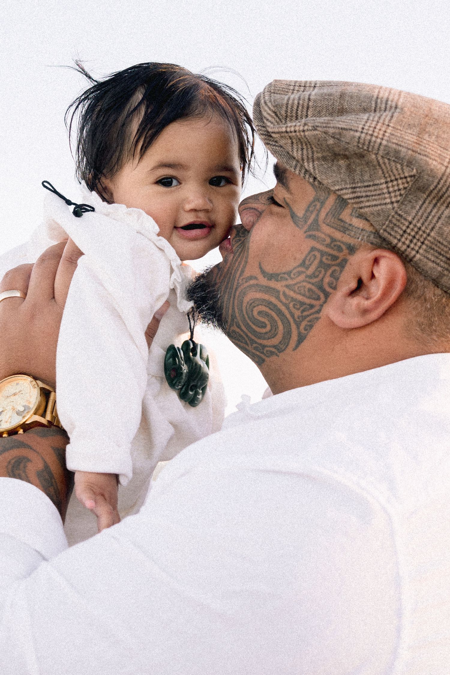 Maori man with facial tattoos kisses baby while holding them up, wearing a green pounamu pendant.