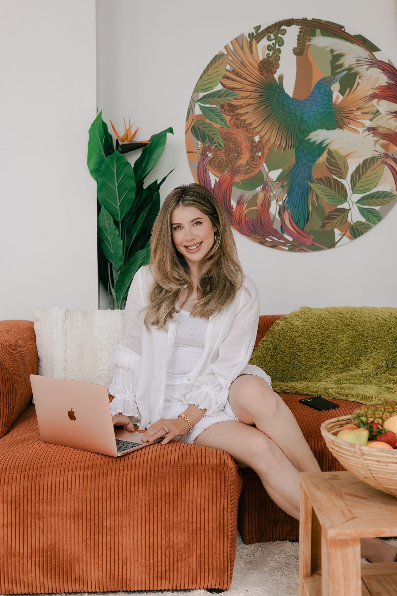 A cozy home office setup with a burnt orange couch, laptop, tropical wall art and indoor plants.