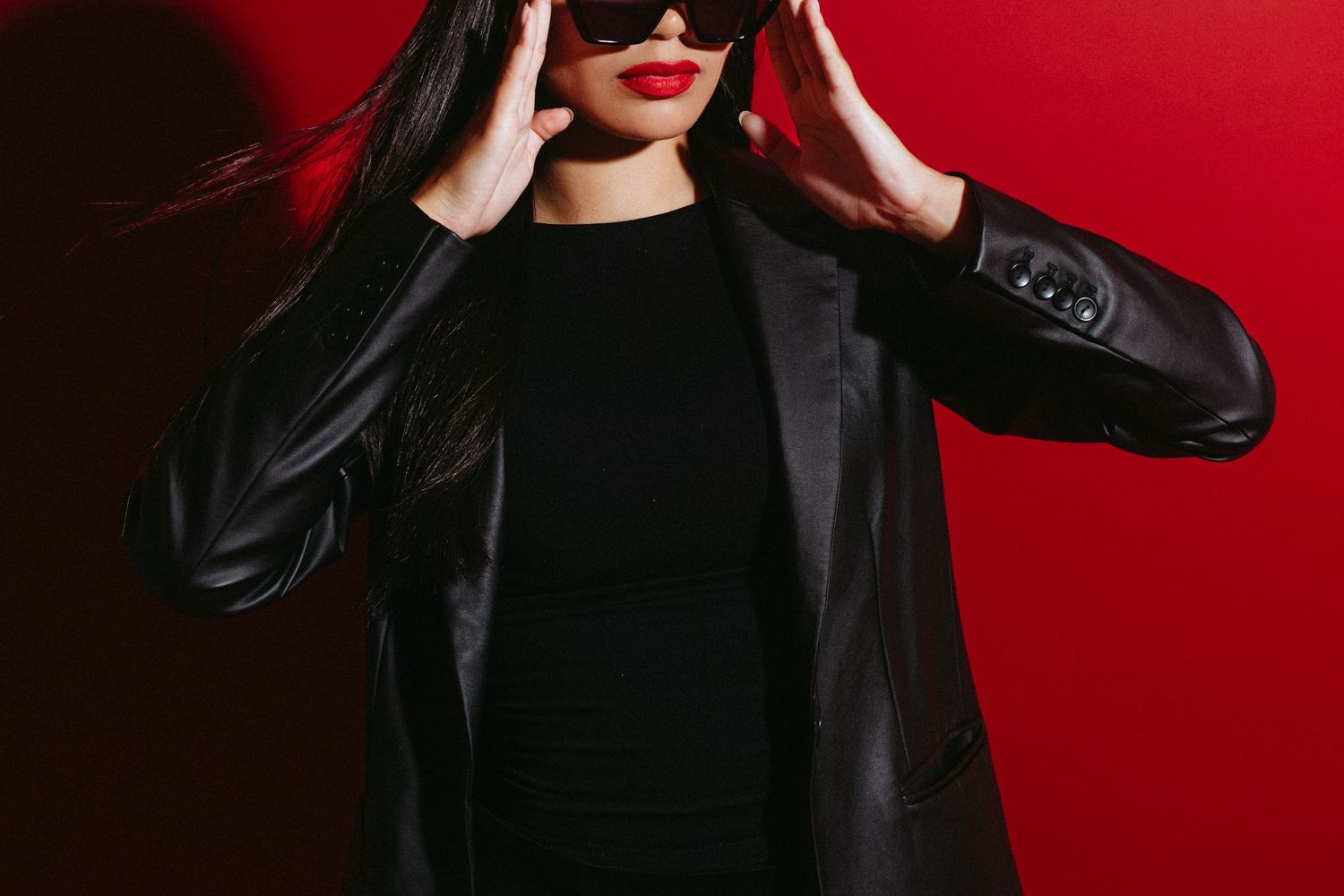 Woman in black leather jacket and red lipstick adjusting sunglasses against a bold red background.