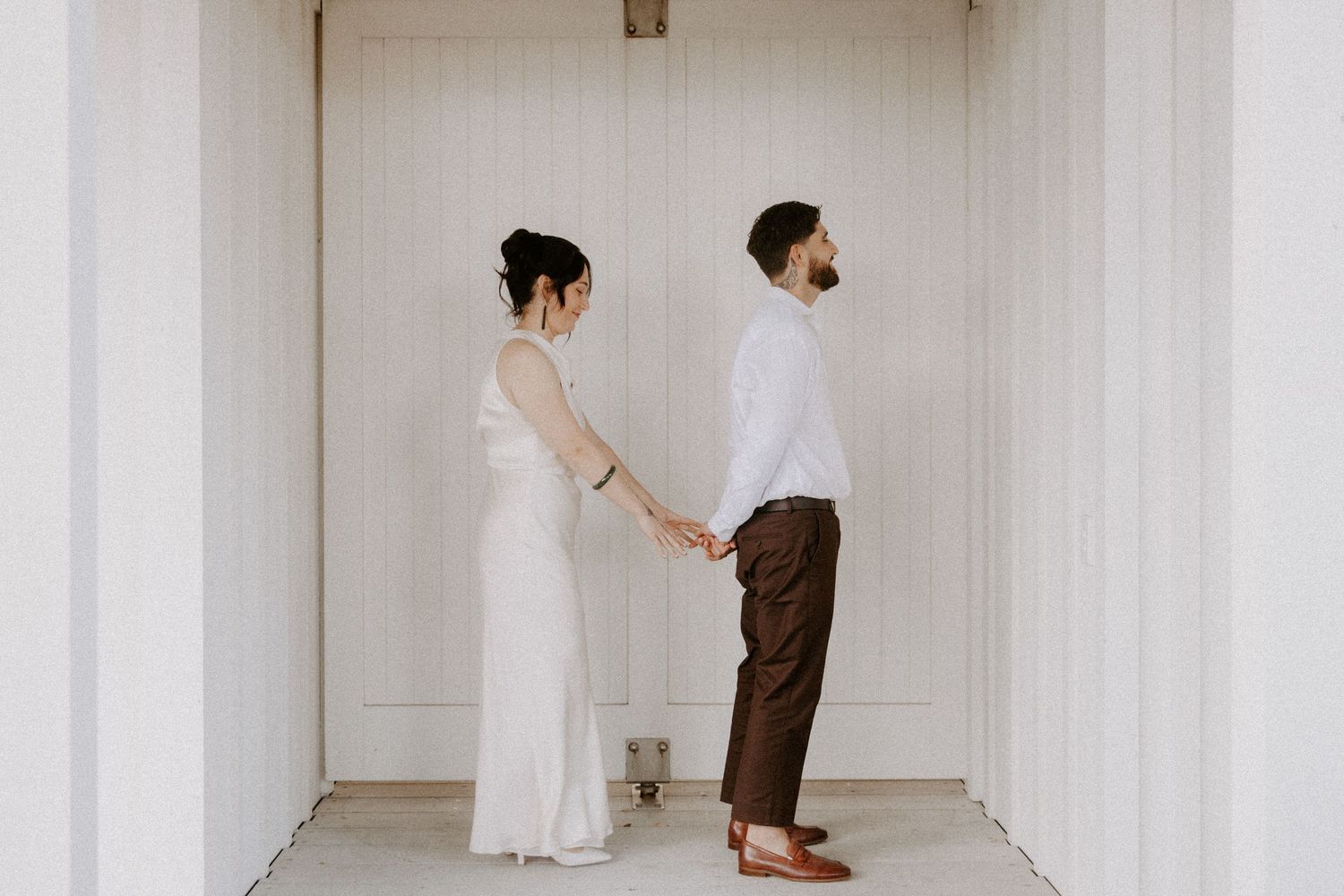 A couple in wedding attire holds hands while standing back to back in a white hallway.