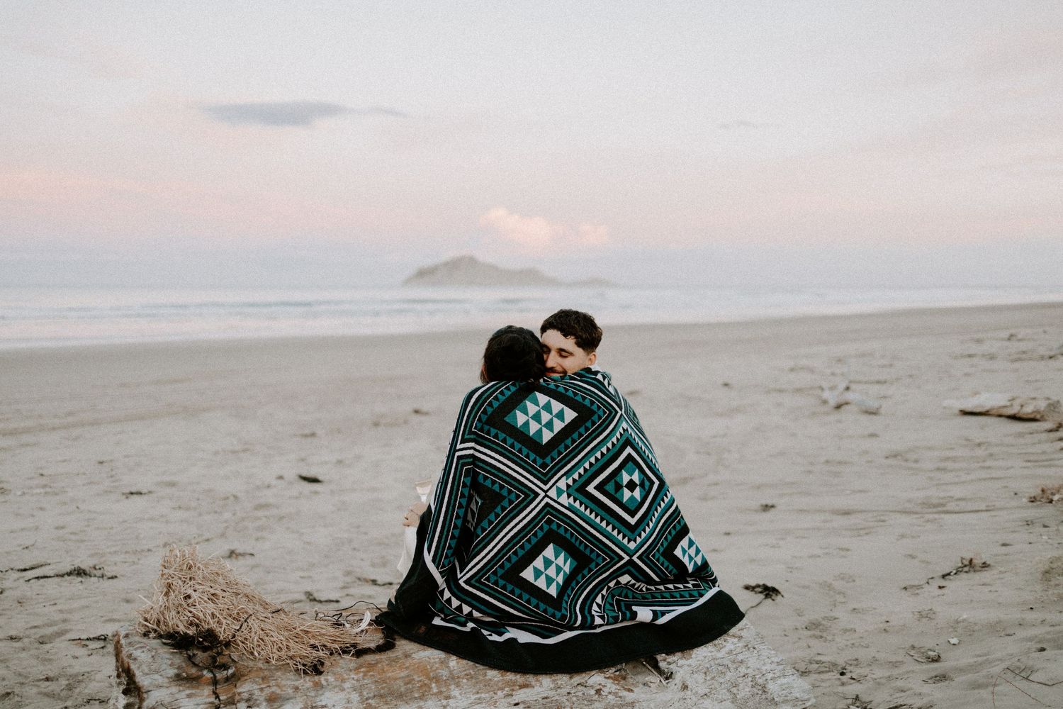 Couple wrapped in patterned blanket sitting on beach during golden hour.