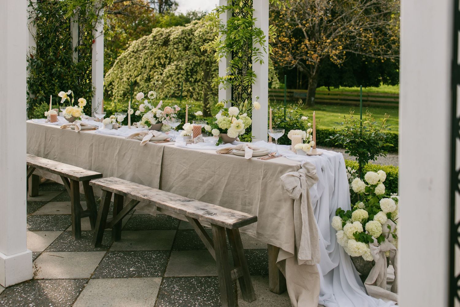 Elegant garden wedding reception table with white linens, rustic benches, and fresh floral arrangements under a covered patio.