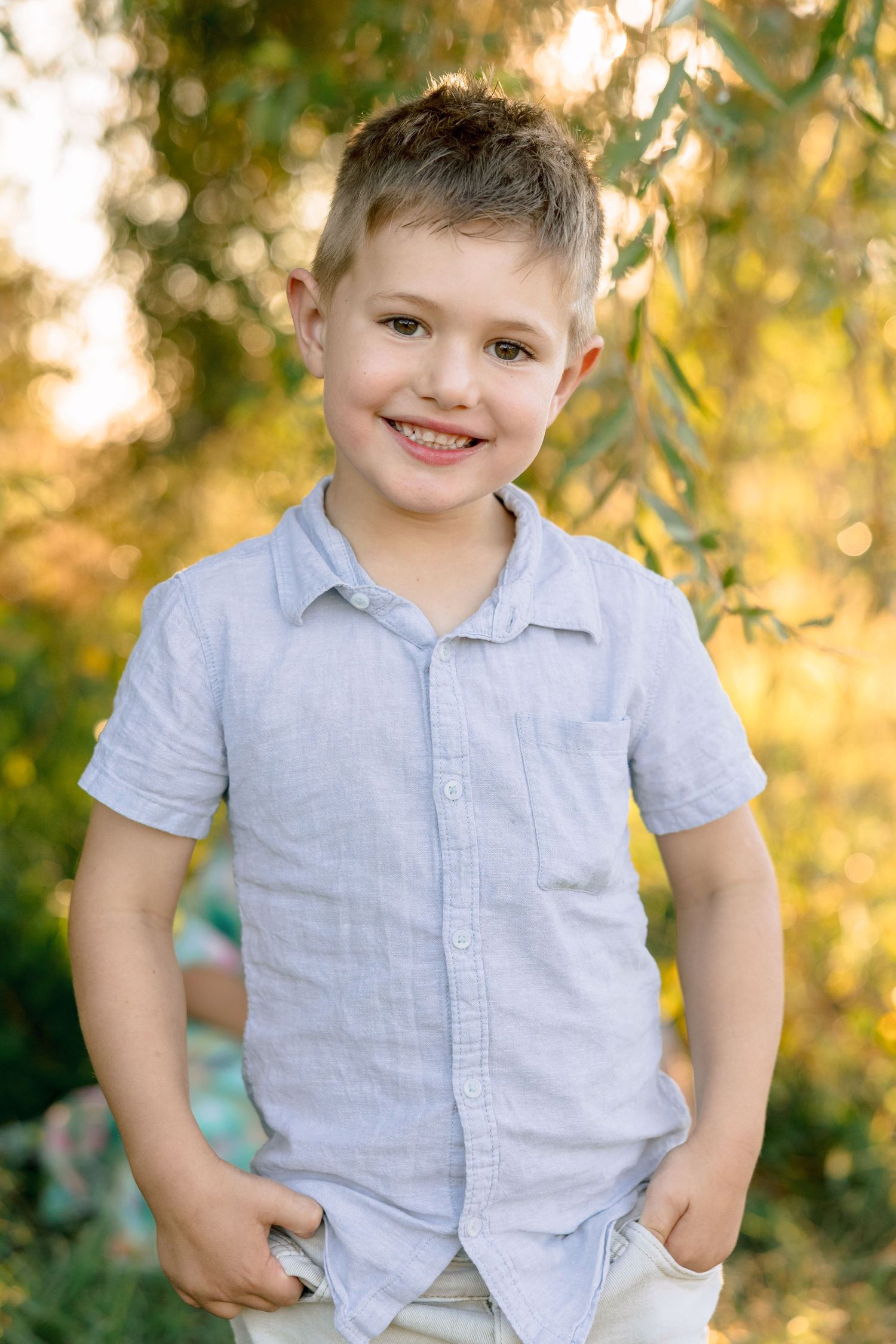 Smiling young boy in a light blue shirt standing outdoors with hands in pockets in golden sunlight.