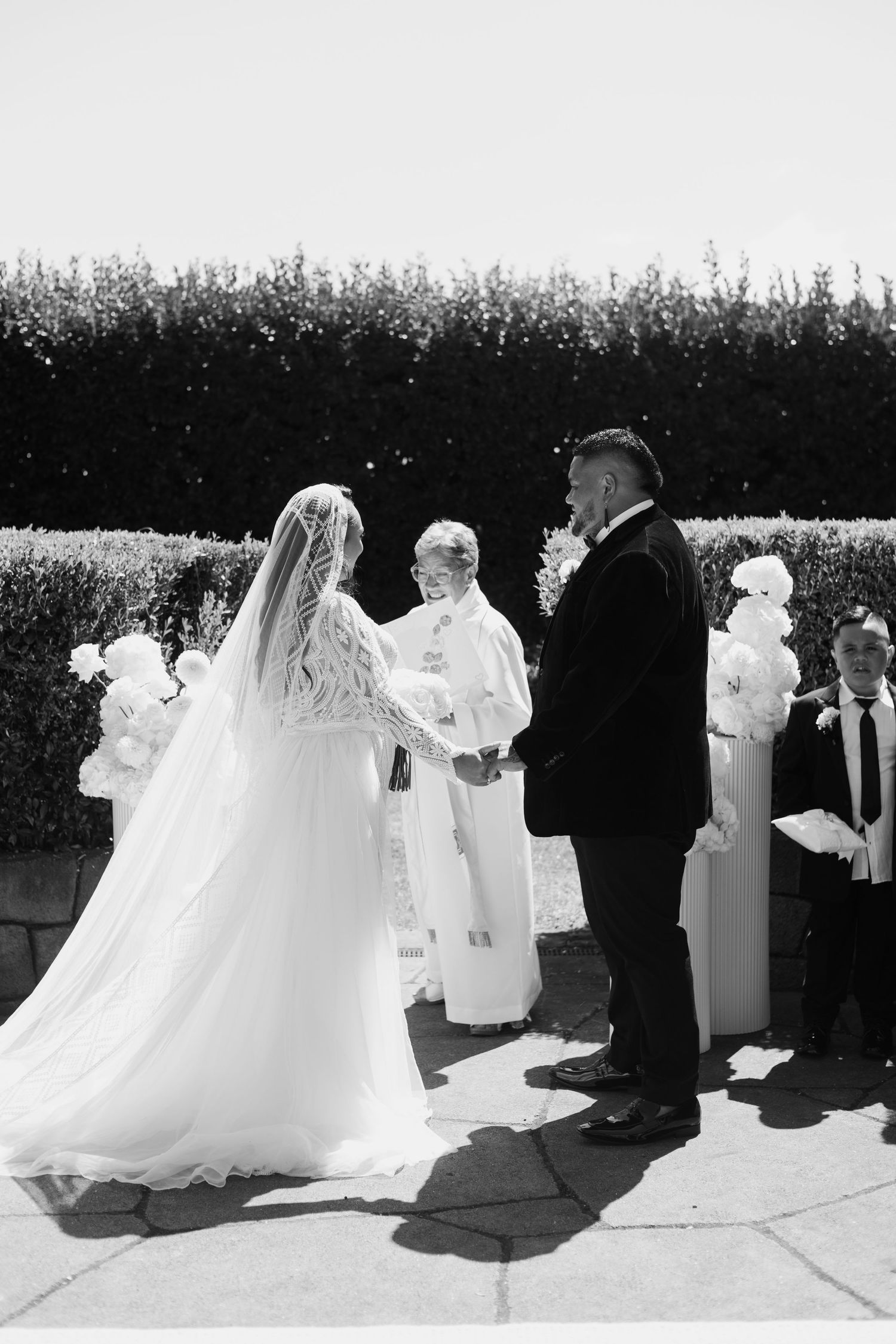 A wedding ceremony taking place outdoors with a couple exchanging vows in front of a hedge backdrop.