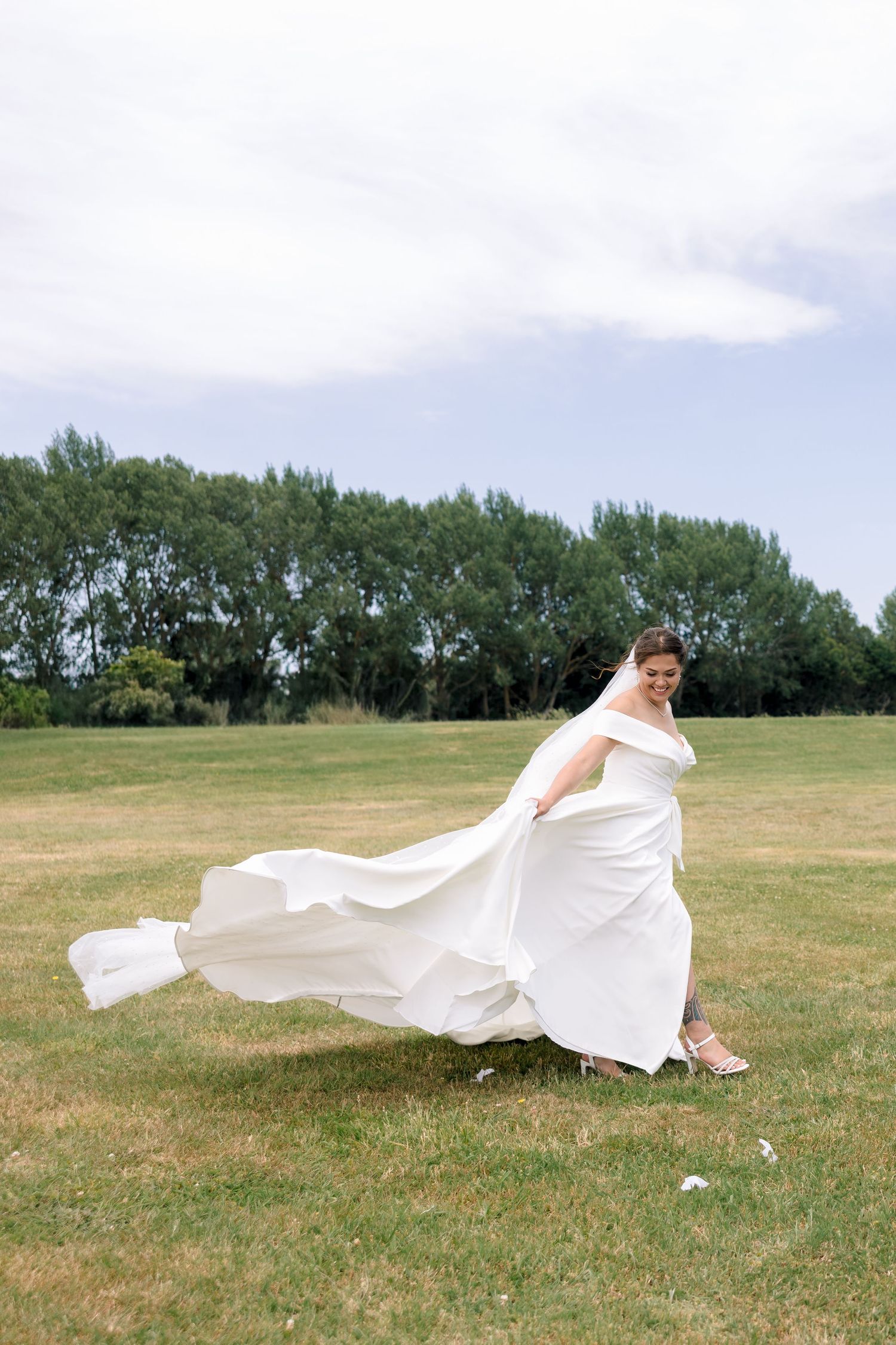 A flowing white dress billows dramatically in the wind across a green grassy field with trees in the background.